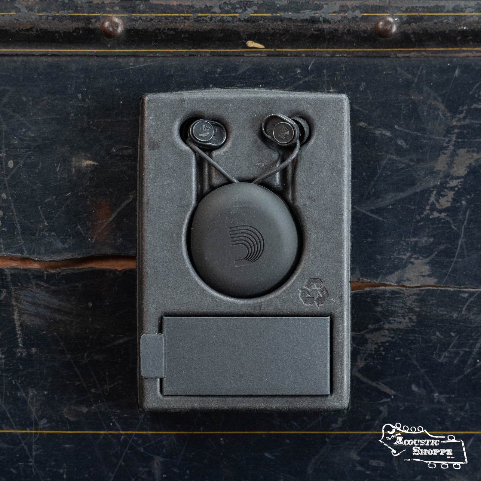 A pair of DAddario PW-DBUDHP-01 Bud Premium Hearing Protection in a black oval case sits in a recycled box on a worn wood surface. Designed for musiciansโ high-fidelity needs, with the Acoustic Shoppe logo at bottom right.