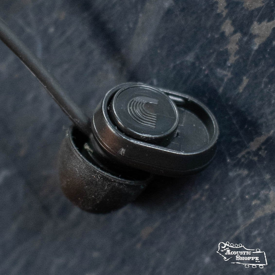 Close-up of the DAddario PW-DBUDHP-01 Bud Premium Hearing Protection earbud, a black wired in-ear model for high-fidelity audio, resting on a dark-textured surface. The Acoustic Shoppe logo is in the bottom right corner.