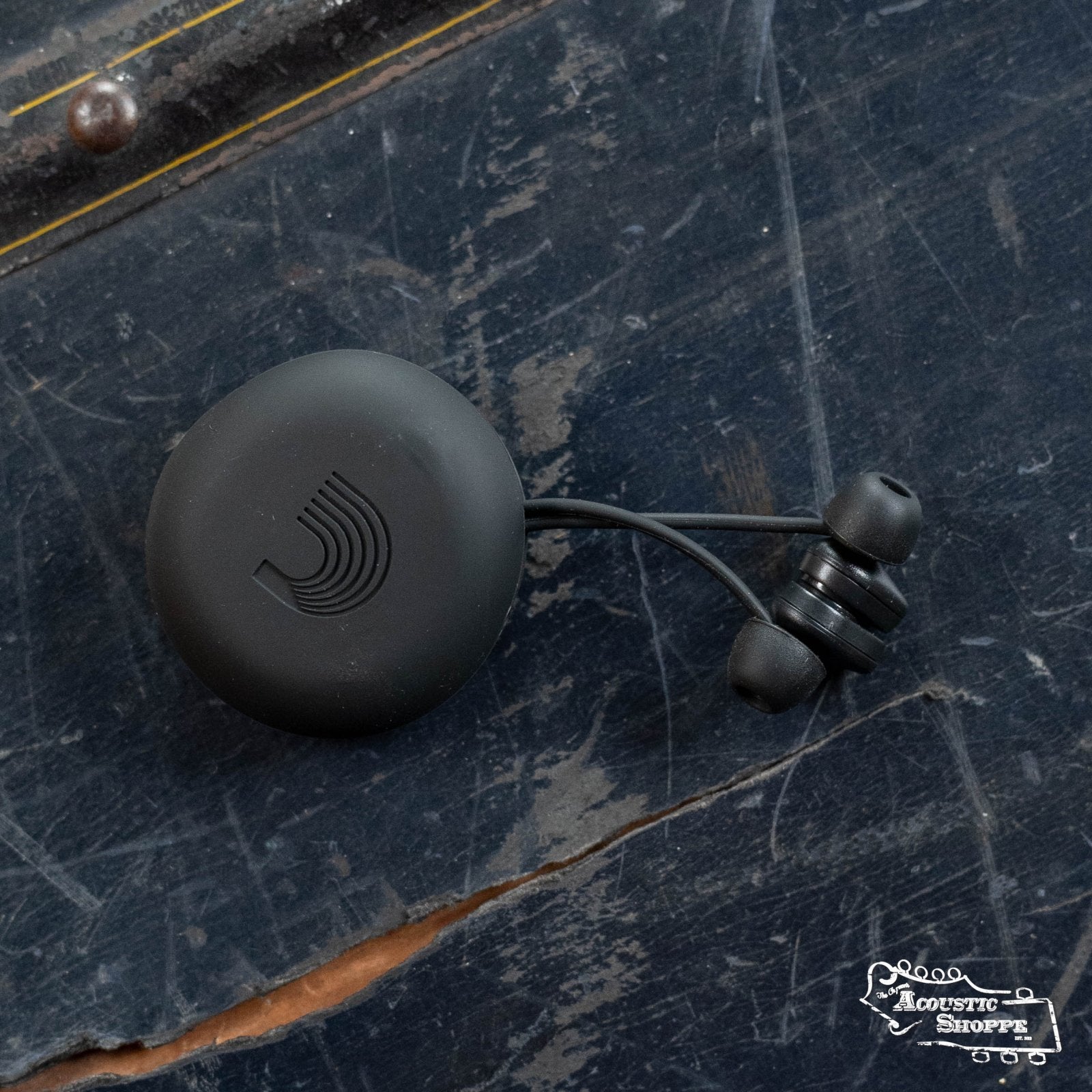 DAddario PW-DBUDHP-01 Bud Premium Hearing Protection by DAddario, wireless black earbuds joined by a short cable, in a round case with an embossed logo, displayed on a dark blue wooden surface. Acoustic Shoppe logo is at the bottom right corner.