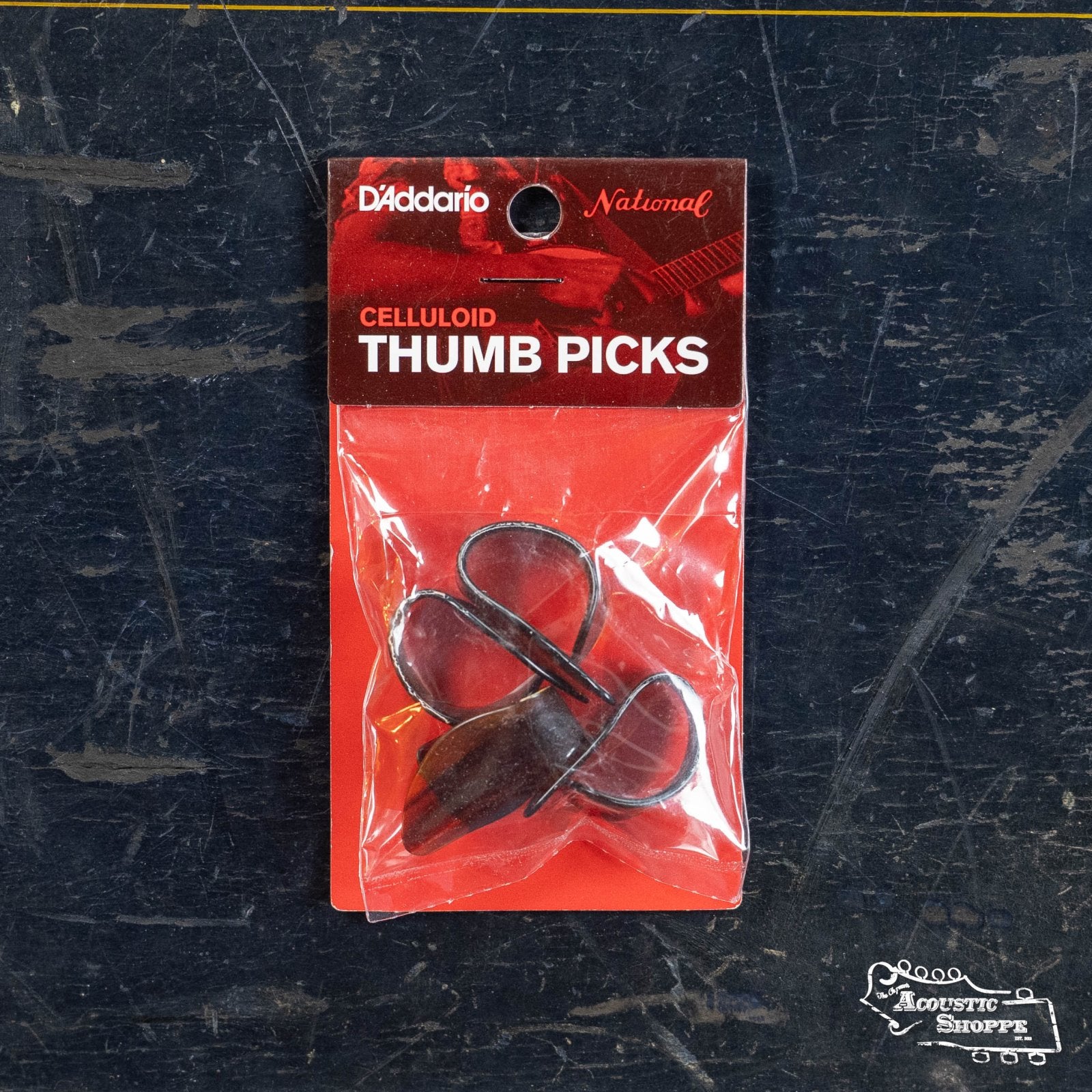 A sealed pack of DAddario NP8T-04 National Celluloid Thumb Picks, Large Tortoiseshell (4-Pack), rests on a dark, scratched surface with The Acoustic Shoppe logo in the lower right corner.