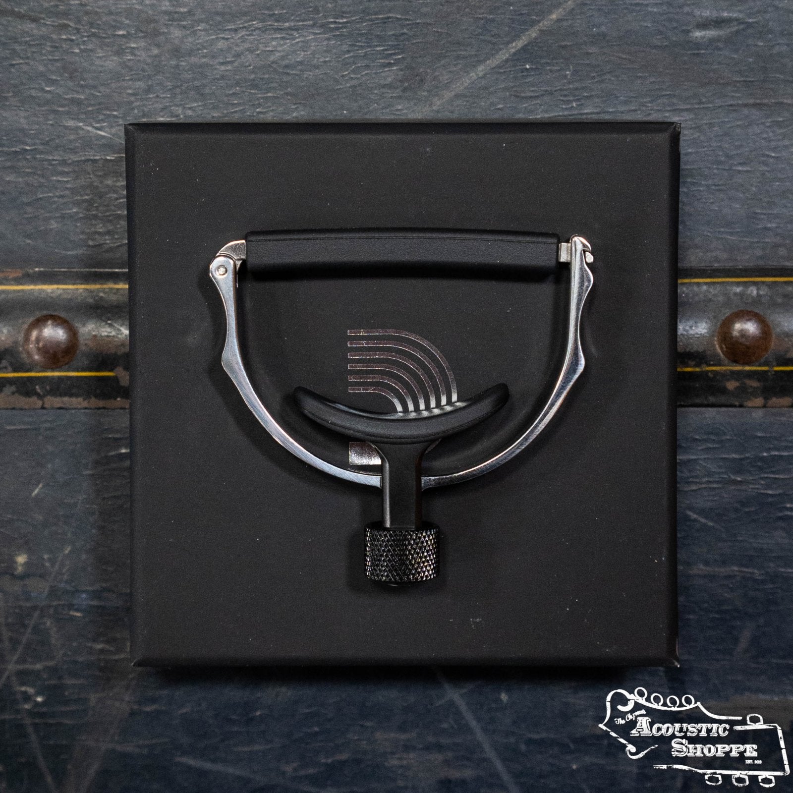 A DAddario PW-CP-18 Guitar Cradle Capo in black stainless steel with a curved design rests on a black box. The backdrop is a textured dark surface with metal rivets, and The Acoustic Shoppe logo appears in white at the bottom right.