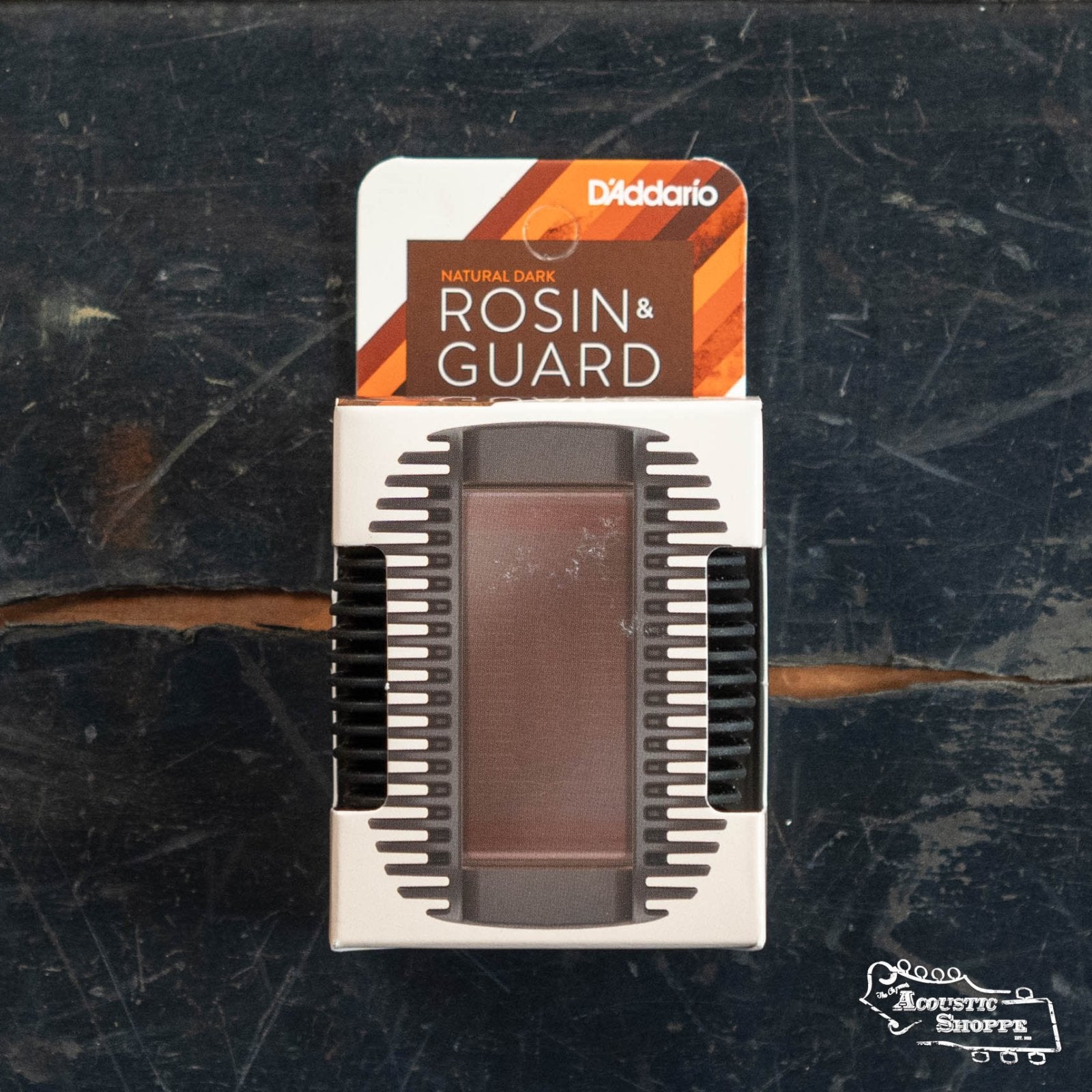 A D'Addario Rosin Guard with Natural Dark Rosin (DRGBK-VR300) sits on a worn wooden surface, its shock-absorbent white and brown packaging revealing premium rosin—an essential accessory for acoustic musicians.