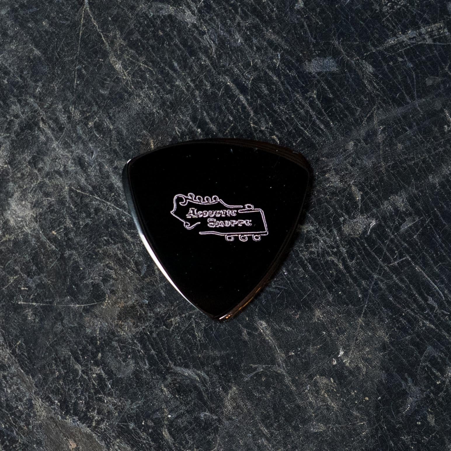 A black ToneSlabs Darth Tone Series Tweener 1.3mm guitar pick with a white ToneSlabs logo rests on a dark, textured surface—an essential accessory for acoustic players and savvy instrument dealers alike.