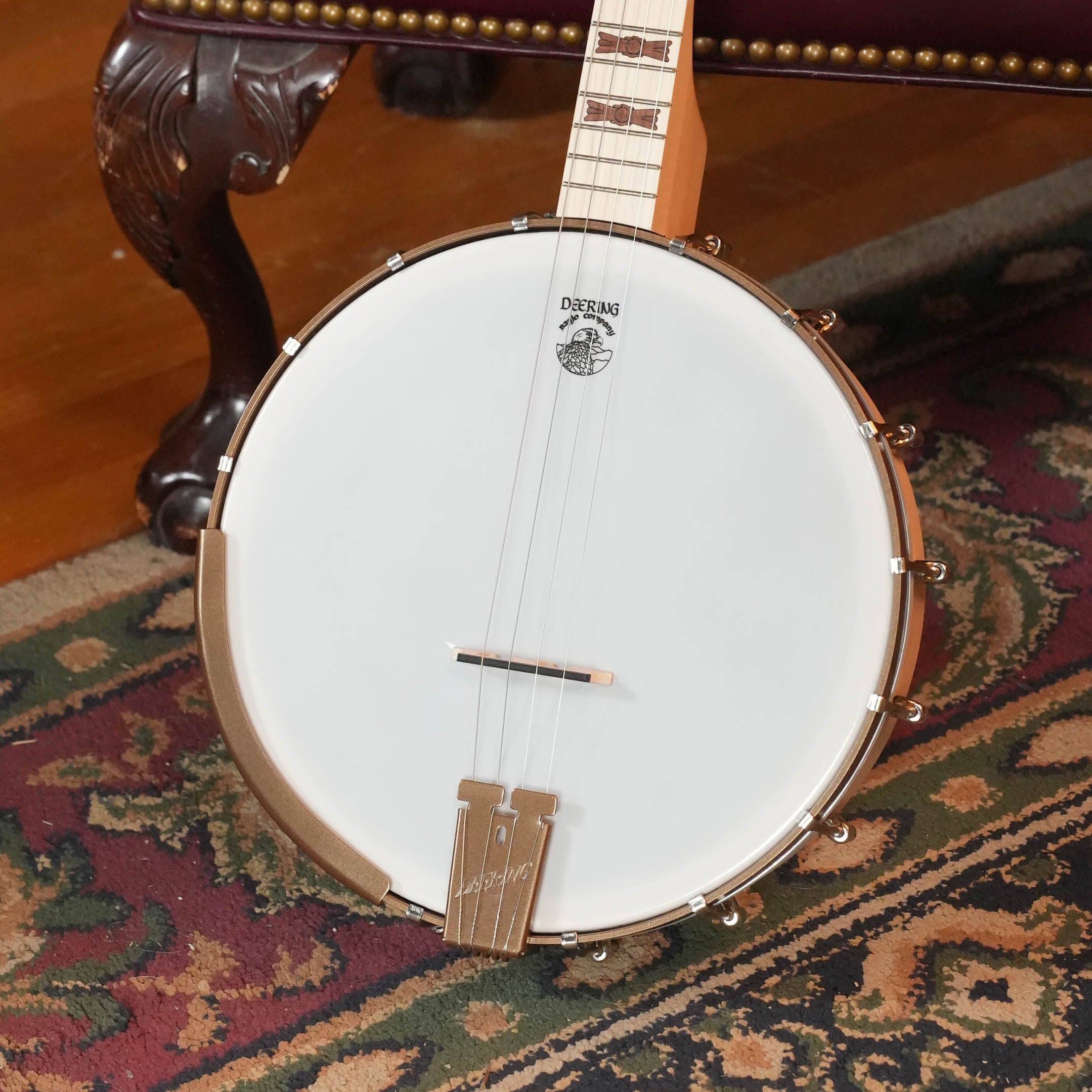 A Deering Banjos Goodtime 19-Fret Tenor Open Back Banjo (#92620) with a white drumhead and maple neck rests on an ornate carpet beside a carved furniture leg accented with brass studs.