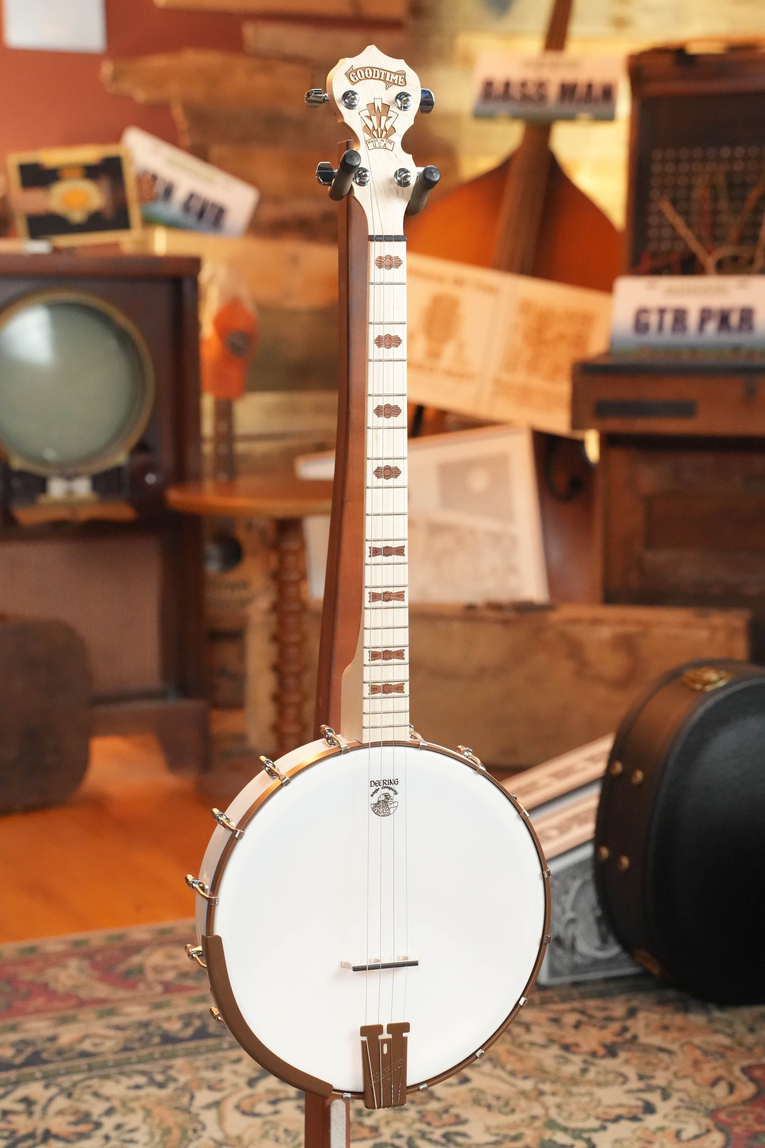 A Deering Banjos Goodtime 19-Fret Tenor Open Back Banjo (#92620) stands upright on a patterned rug, encircled by vintage musical gear, instruments, and sheet music in a warmly lit room.