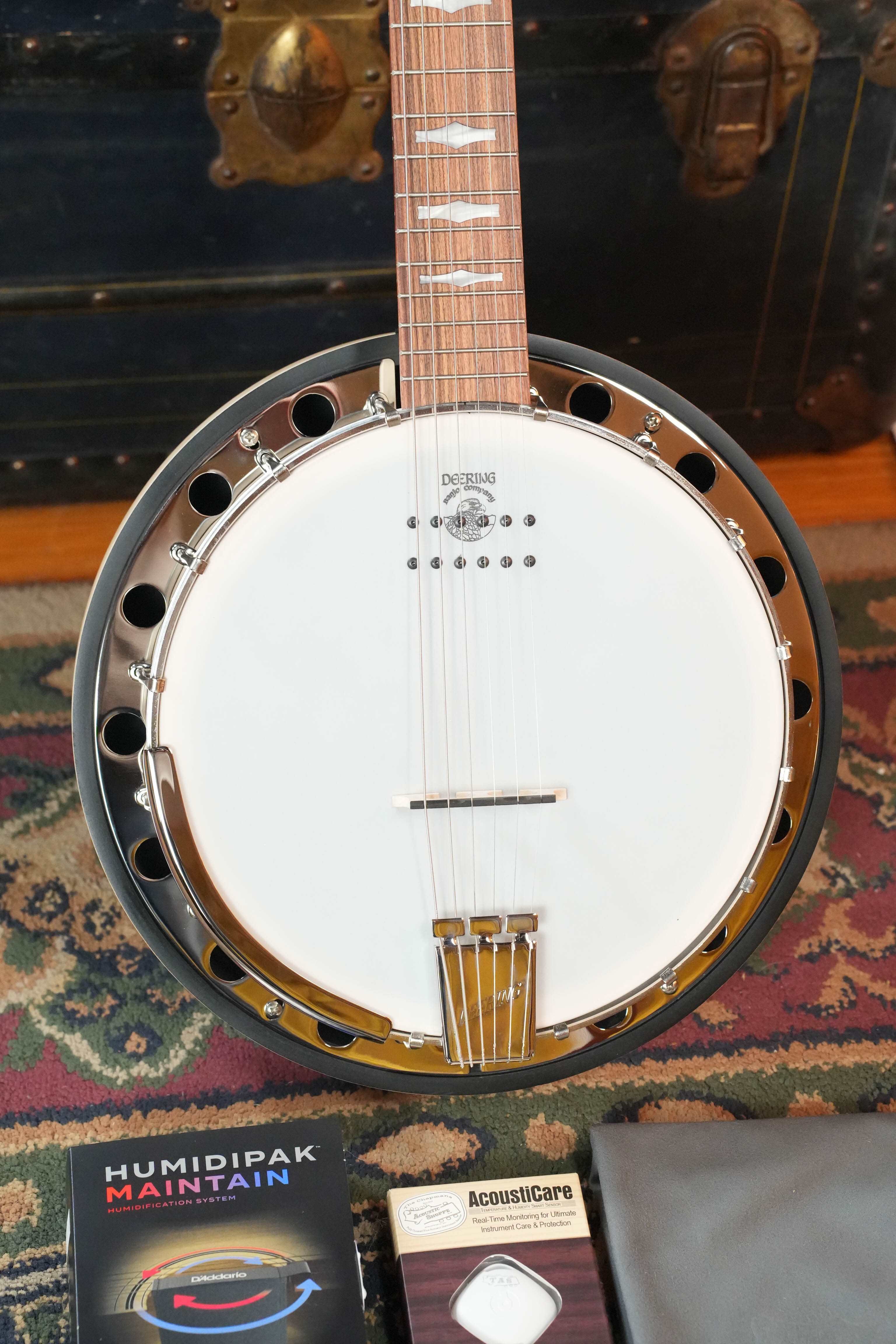 A Deering Banjos Goodtime 6 R Steel String Banjo with Kavanjo Pickup rests on a patterned rug. Nearby are a Humidipak Maintain package, an AcoustiCare item, and a gray cloth. A vintage trunk sits in the background.