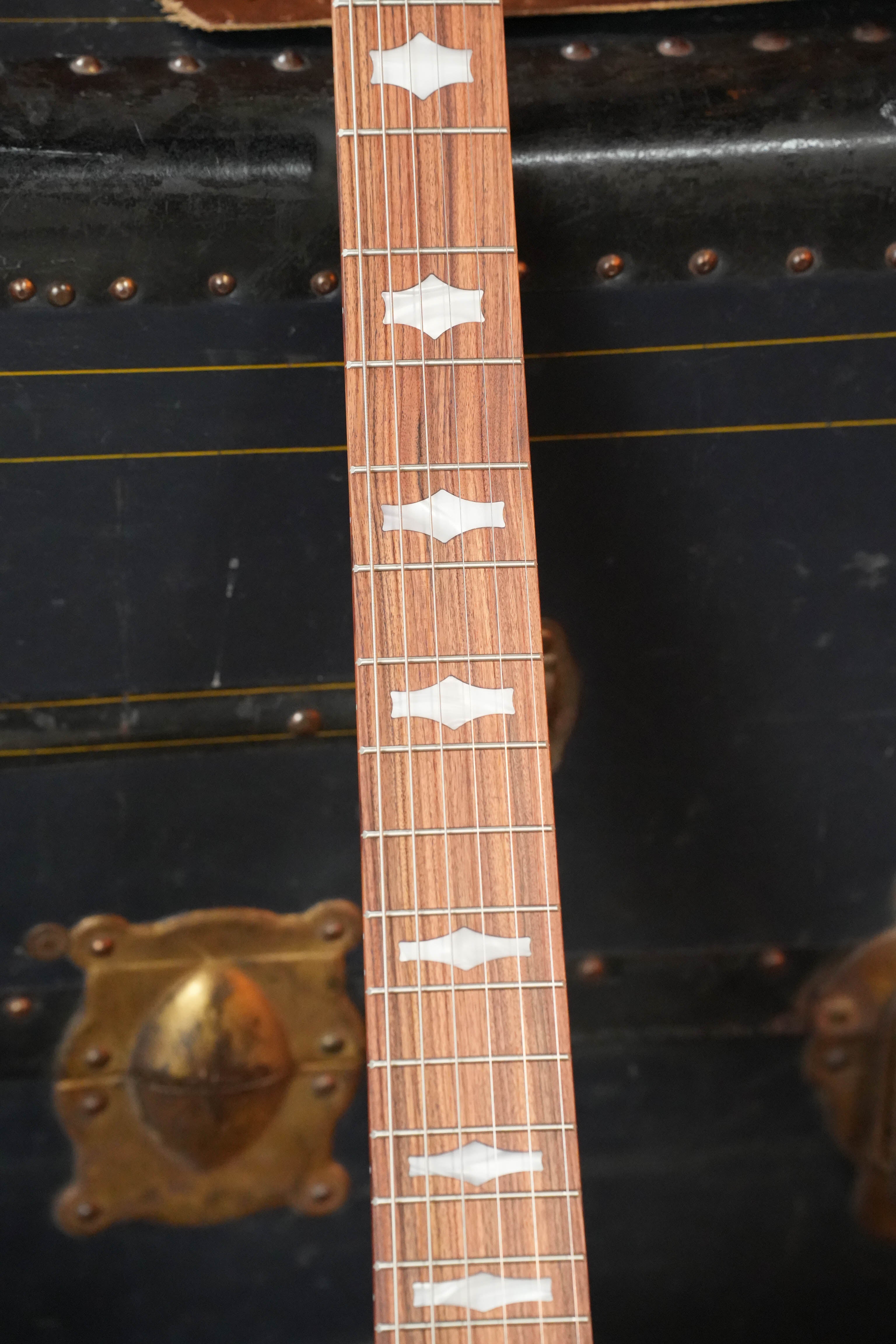 A close-up of the Deering Goodtime 6 R Steel String Banjo w/Kavanjo Pickup by Deering Banjos shows unique white fretboard inlays, set on an old black trunk with brass hardware—ideal for your next acoustic jam.