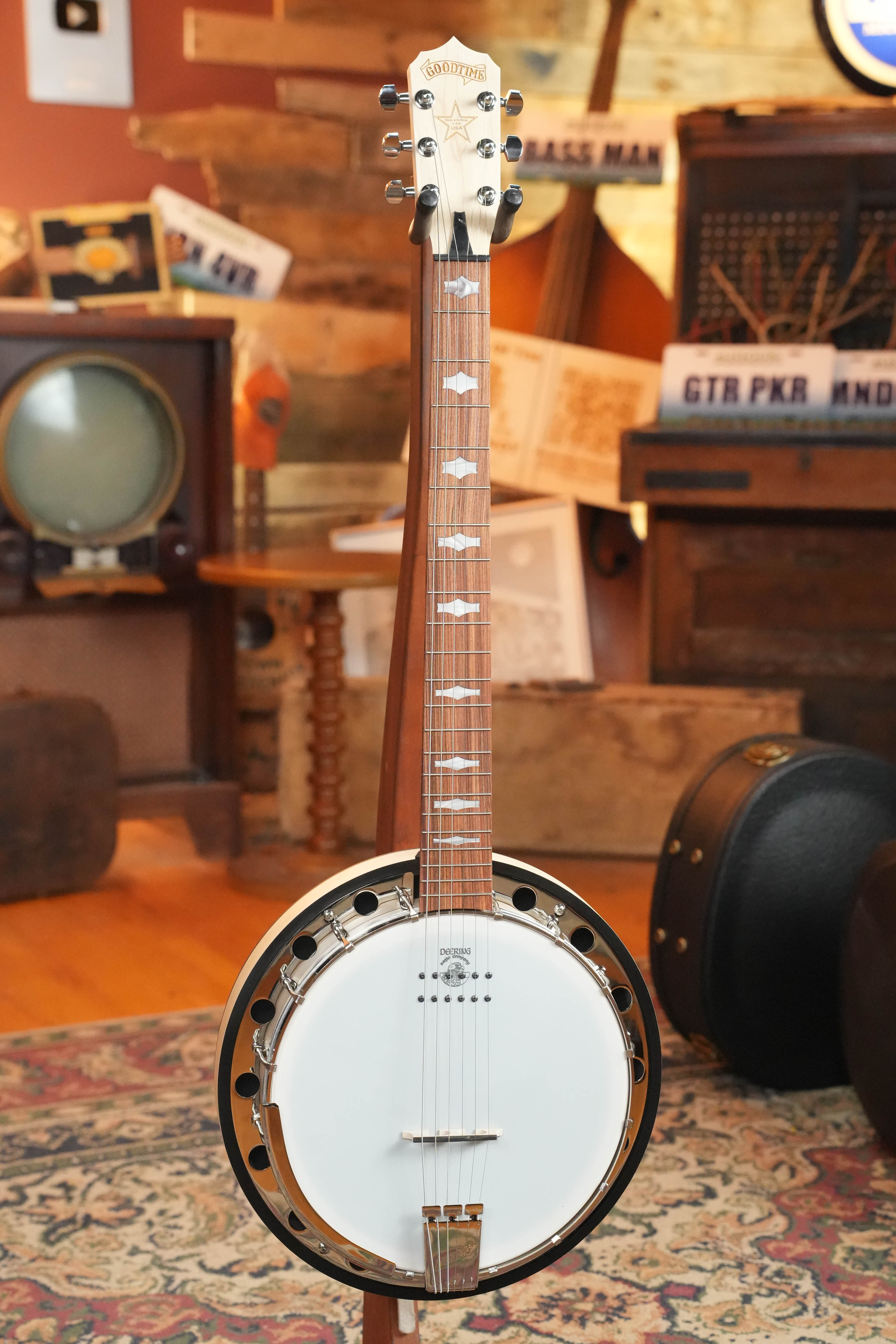 The Deering Goodtime 6 R Steel String Banjo W/Kavanjo Pickup #2620 by Deering Banjos stands upright in a room filled with vintage musical and television equipment.