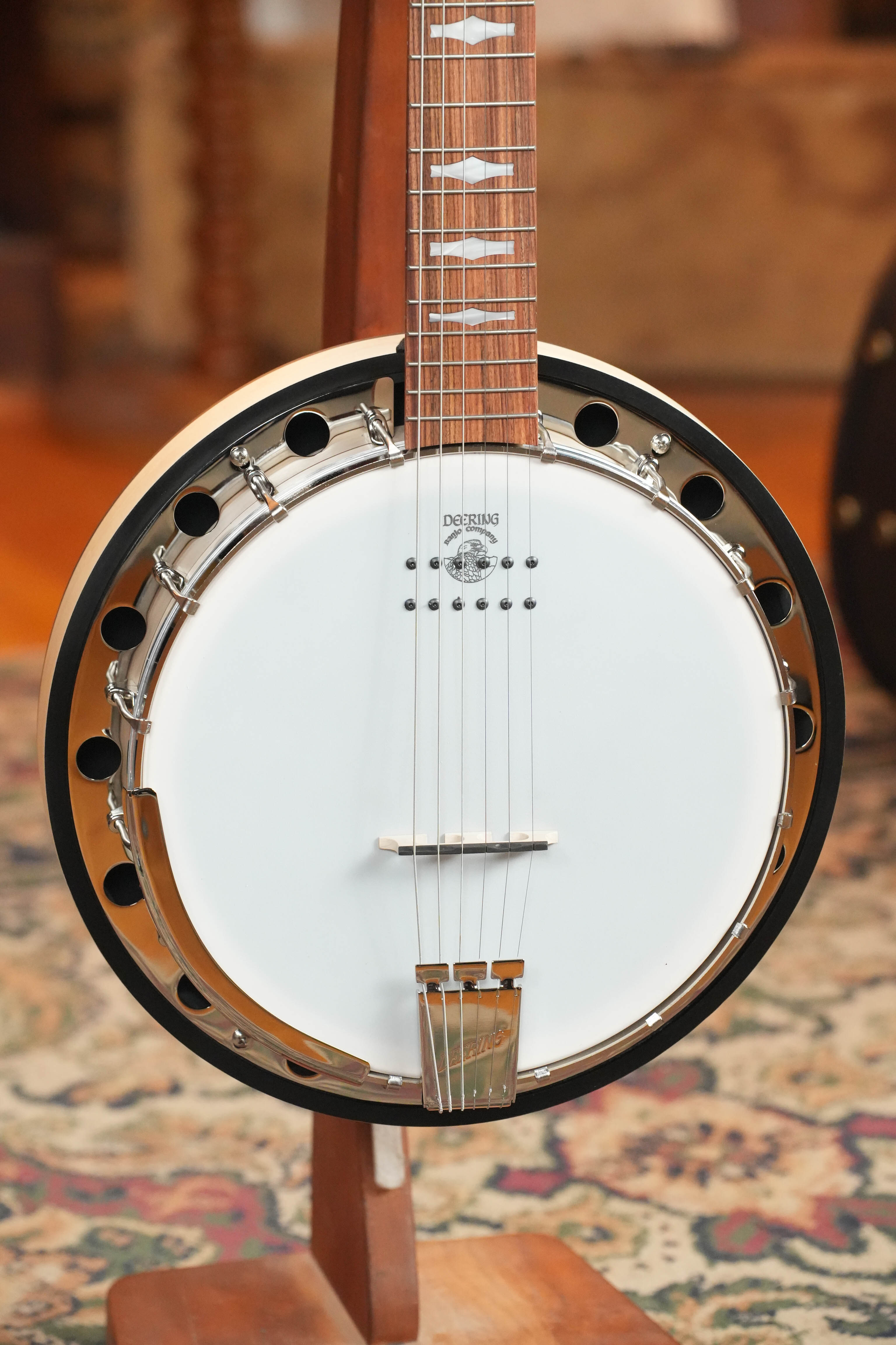 A close-up of the Deering Banjos Goodtime 6 R Steel String Banjo w/Kavanjo Pickup #2620 displays its round white drumhead, metallic rim with holes, and strings on a wooden stand over a patterned rug background.