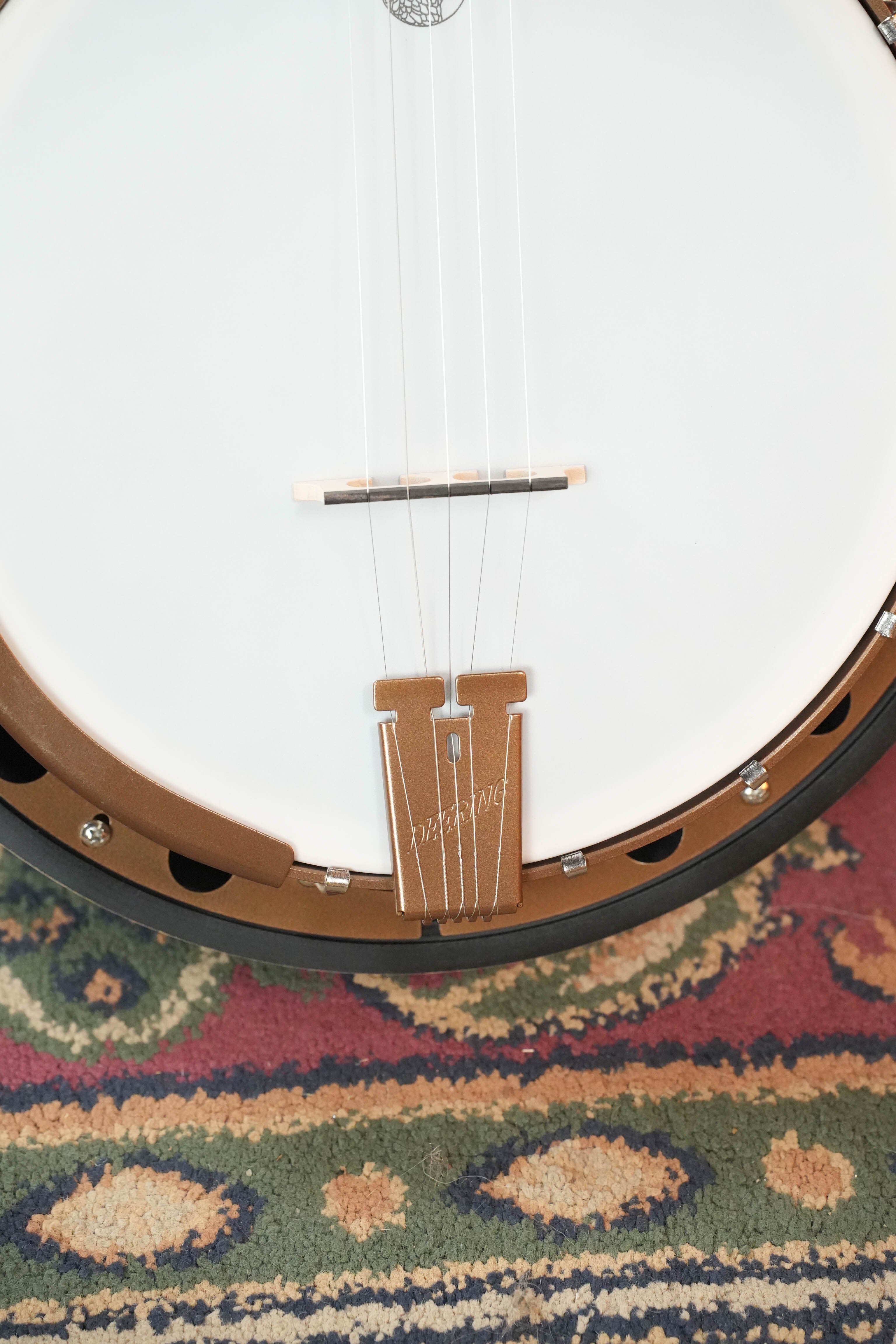 Close-up of a Deering Goodtime Two 5 String Resonator Banjo #2615 by Deering Banjos, showing its body, tailpiece, strings, bridge, and part of the banjo head resting on a colorful patterned rug.