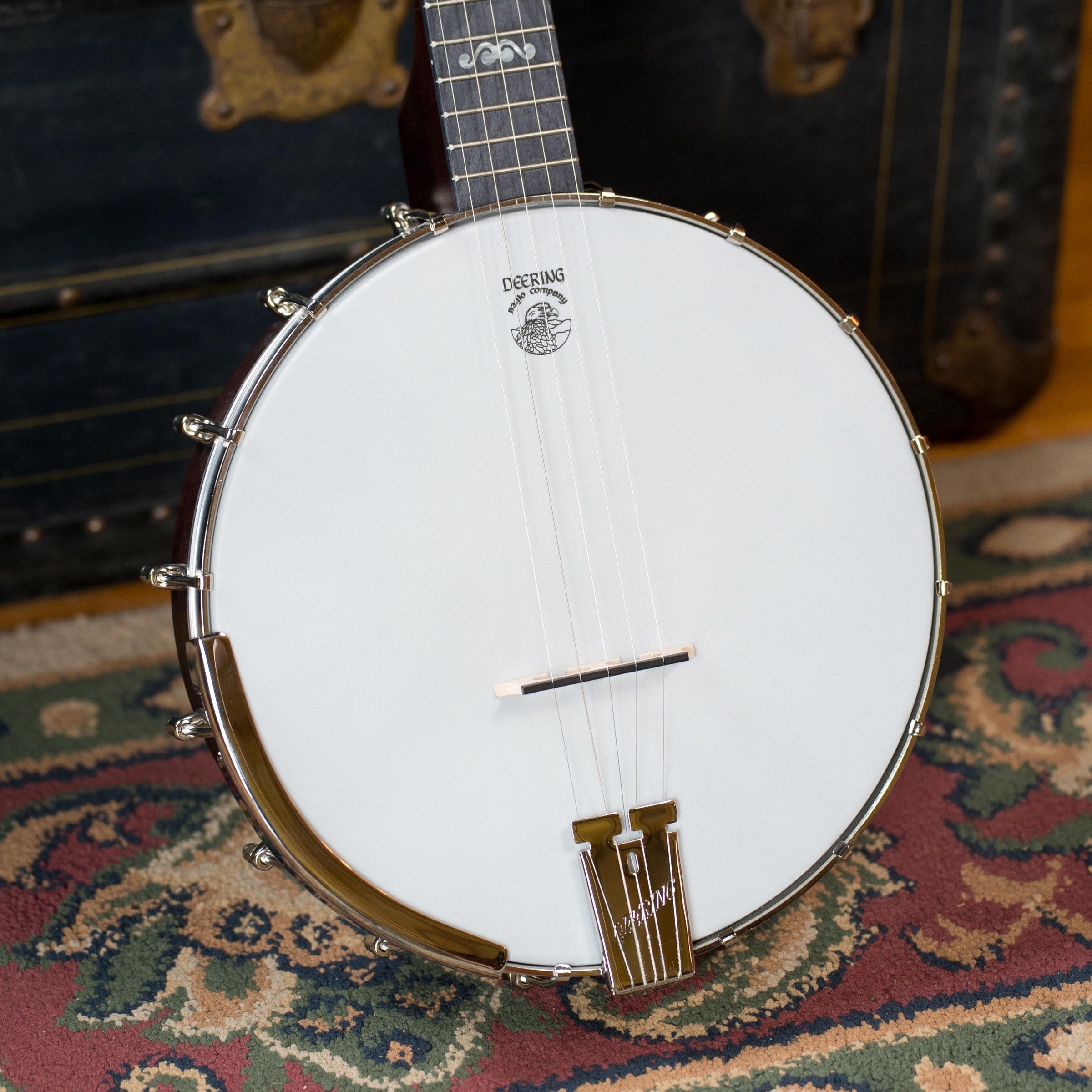 A close-up of the Deering Banjos Artisan Goodtime 5-String Openback Banjo #6251 resting on an ornate patterned rug, with its white drumhead, metal hardware, and wooden neck visible. A vintage trunk appears in the background.