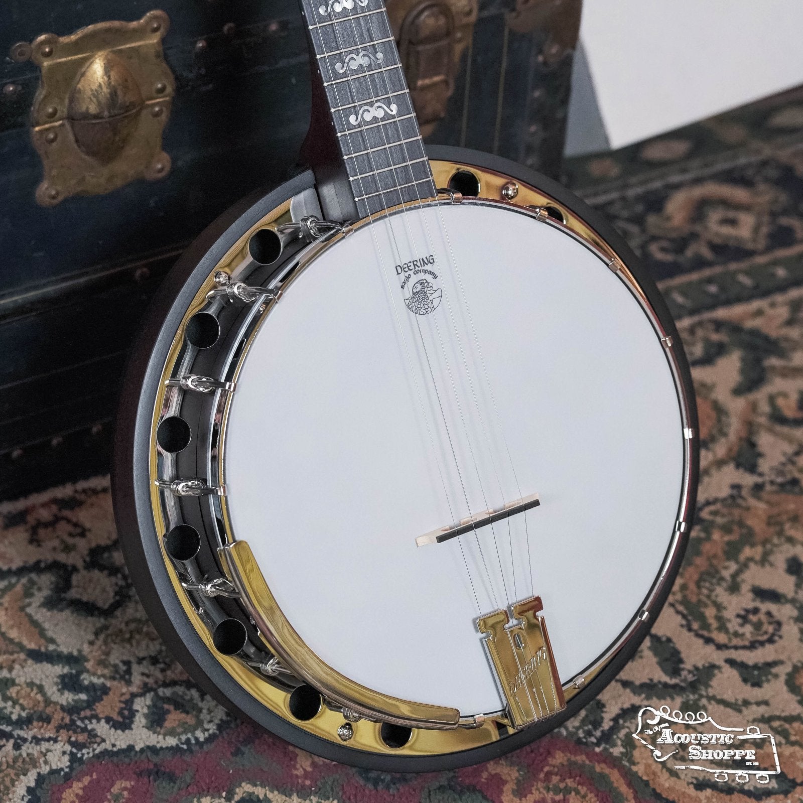 A close-up of the Deering Artisan Goodtime Two 5-String Banjo with Resonator #8977 by Deering Banjos, featuring a white drumhead, gold hardware, and neck inlays, rests on a patterned rug near a vintage trunk. The Acoustic Shoppe logo appears bottom right.