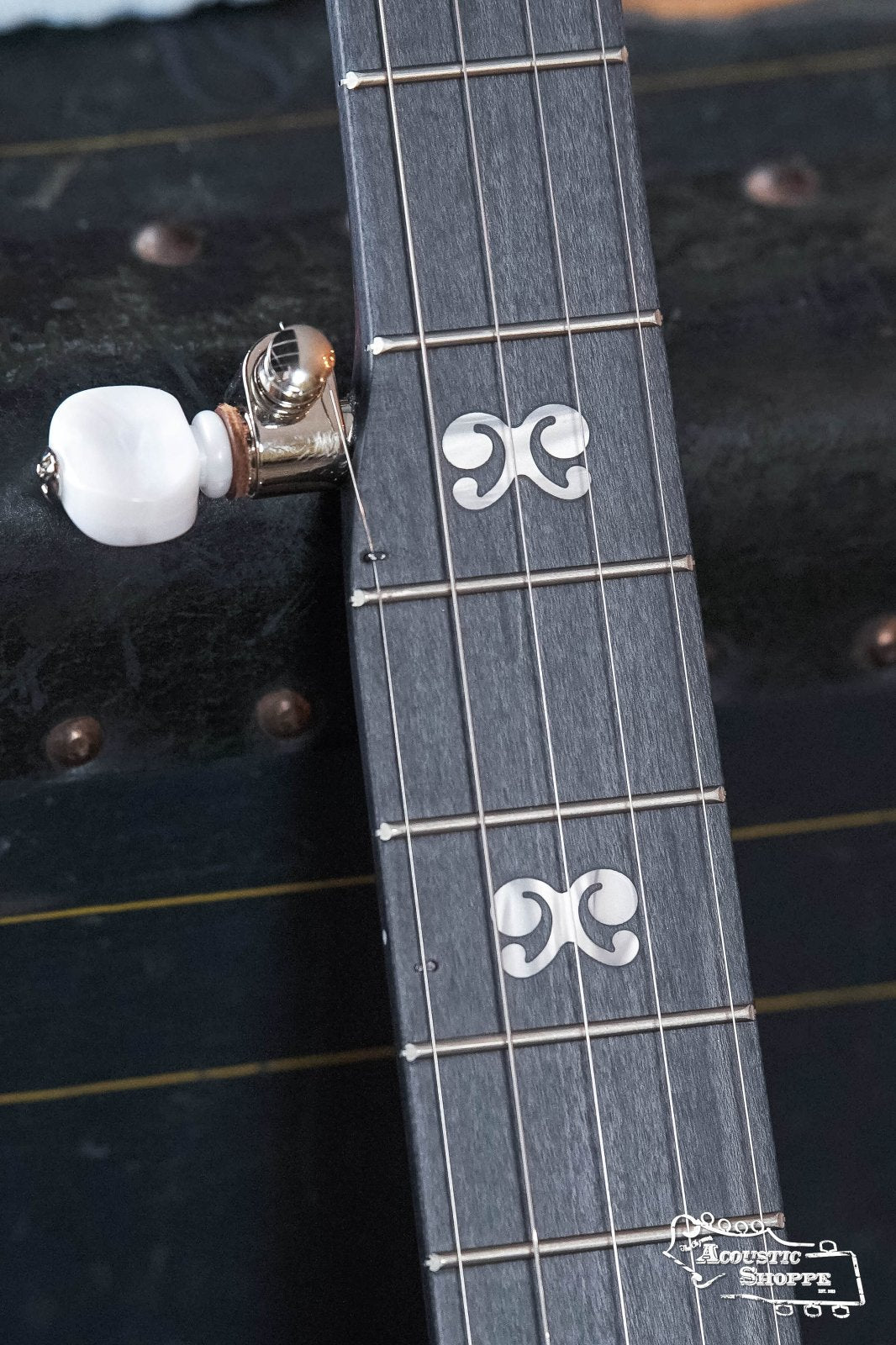 Close-up of a Deering Artisan Goodtime Two 5-String Banjo with Resonator #8977 by Deering Banjos, highlighting the decorative fretboard inlays and white tuning peg, with a vintage trunk visible in the background.