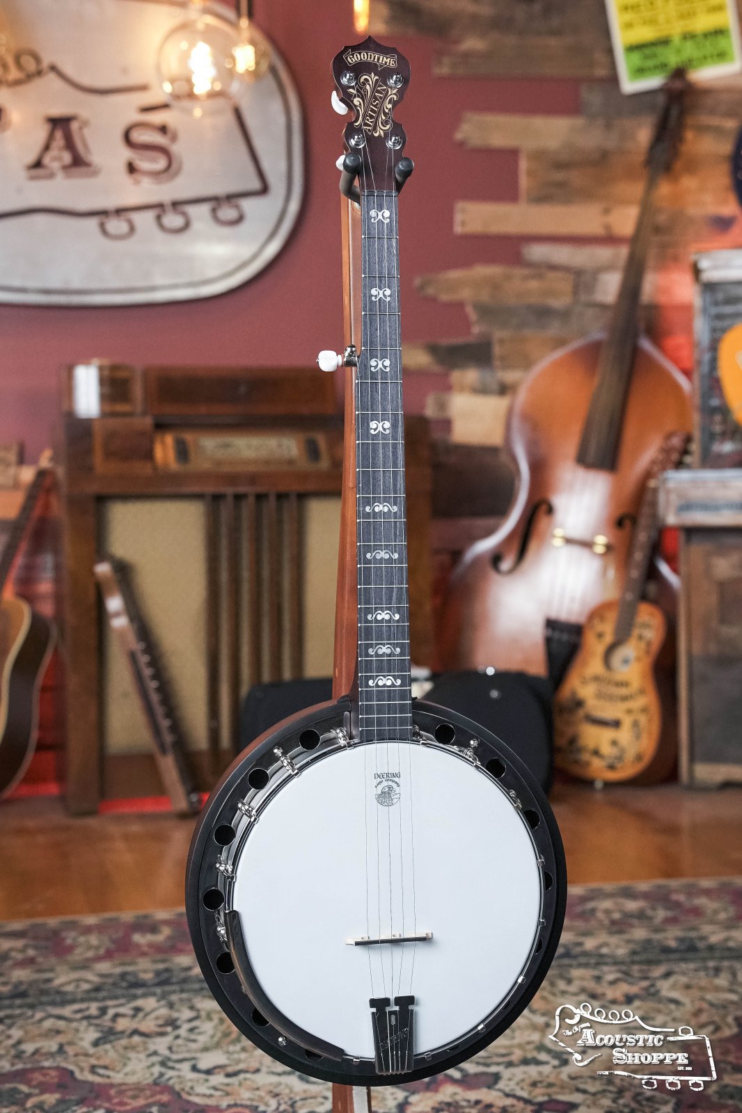 A Deering Artisan Goodtime Two 5-String Banjo with Resonator (#8977) by Deering Banjos stands in The Acoustic Shoppe, surrounded by string instruments and vintage decor. The shop’s logo appears in the corner.