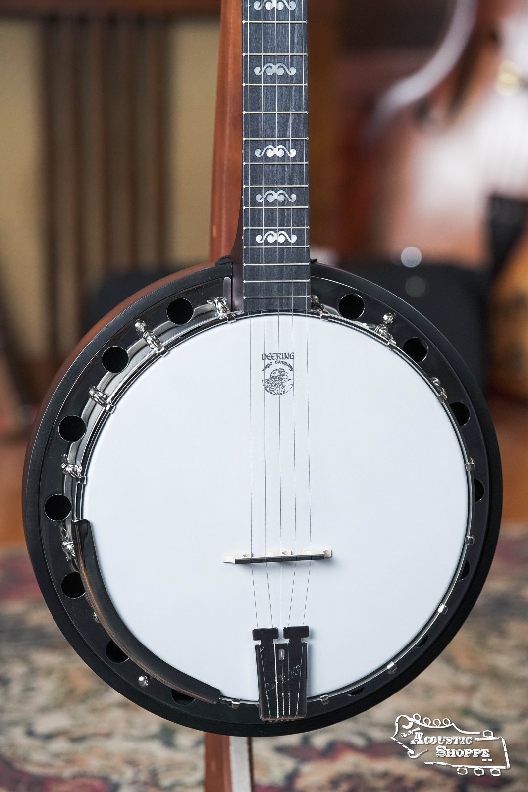 Close-up of a black Deering Banjos Artisan Goodtime Two 5-String Banjo with Resonator (#8977) on a stand, featuring its round white body, strings, and fretboard. The “Acoustic Shoppe” logo appears in the lower right against a softly blurred background.