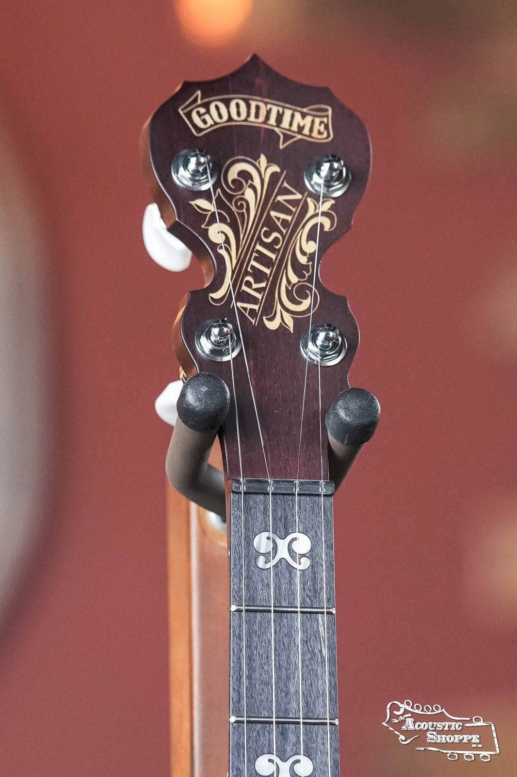 Close-up of the Deering Artisan Goodtime Two 5-String Banjo with Resonator (#8977) headstock by Deering Banjos, featuring ornate branding, partial fretboard and tuners, a blurred reddish background, and Acoustic Shoppe watermark.