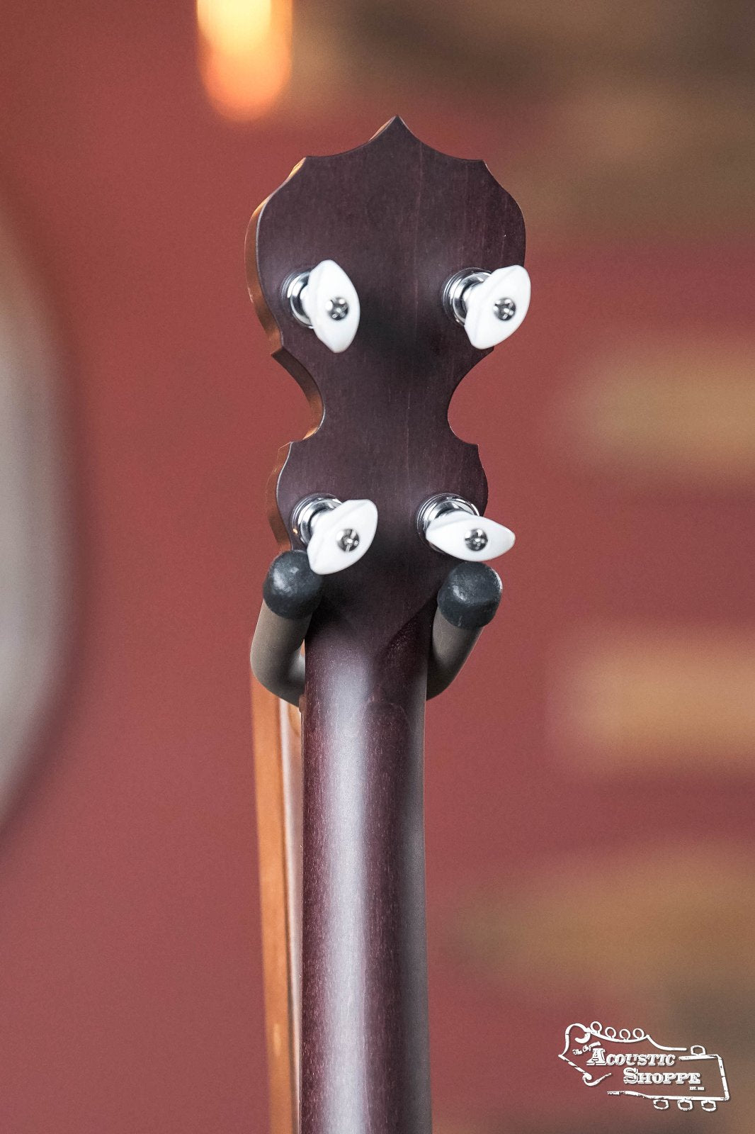 Close-up of the back of a Deering Artisan Goodtime Two 5-String Banjo with Resonator (#8977) by Deering Banjos, showing four tuning pegs against a blurred reddish-brown background. The Acoustic Shoppe logo is in the bottom right corner.