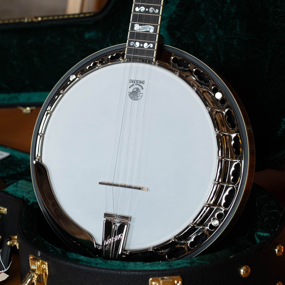 A close-up of a Deering Banjos Calico 5 String Curly Maple Resonator Banjo w/Spikes Installed #AX45 in its open black case with gold latches, featuring a white drumhead and metallic hardware against a dark green interior.
