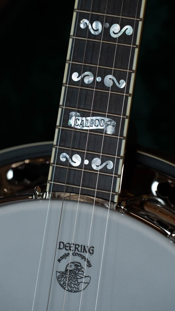 Close-up of a Deering Calico 5 String Curly Maple Resonator Banjo w/Spikes Installed (#AX45) by Deering Banjos, showing decorative inlays on the neck and CALICO on the fretboard, with the Deering logo on the head.