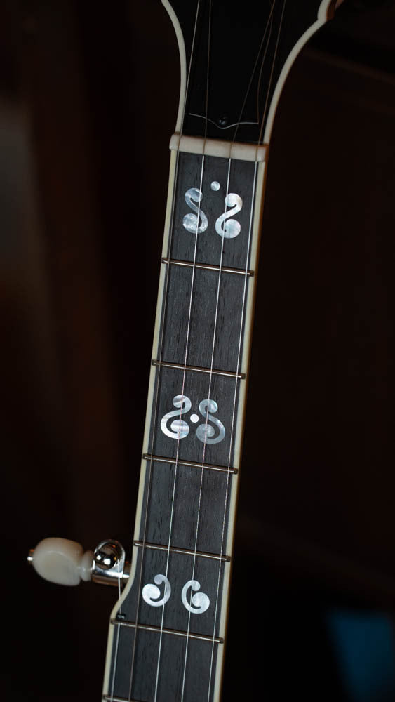 A close-up of the Deering Banjos Calico 5 String Curly Maple Resonator Banjo w/Spikes Installed (#AX45) displays unique music note inlays on a dark fretboard and cream binding, highlighting this professional Deering models craftsmanship.