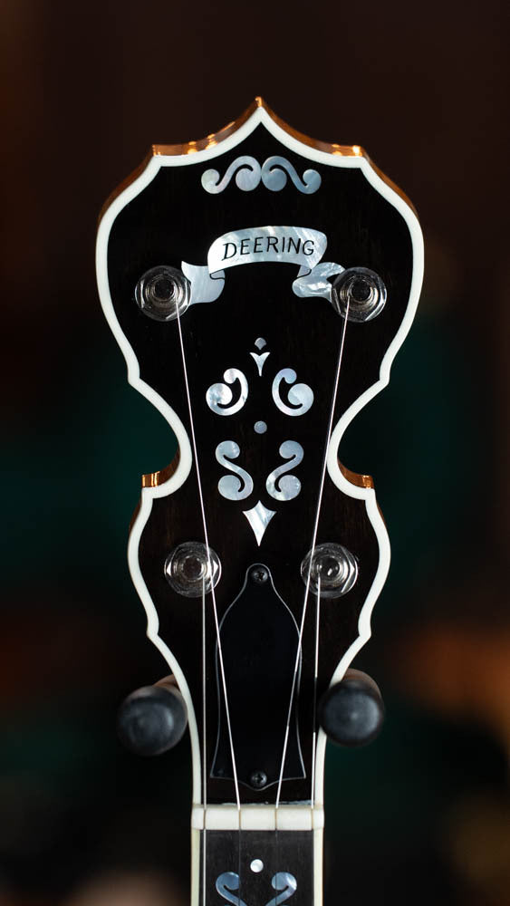 Close-up of a Deering Banjos Calico 5 String Curly Maple Resonator Banjo w/Spikes Installed #AX45 headstock, showing ornate white inlays, banner-shaped Deering, plus tuning pegs and strings that highlight its professional craftsmanship.