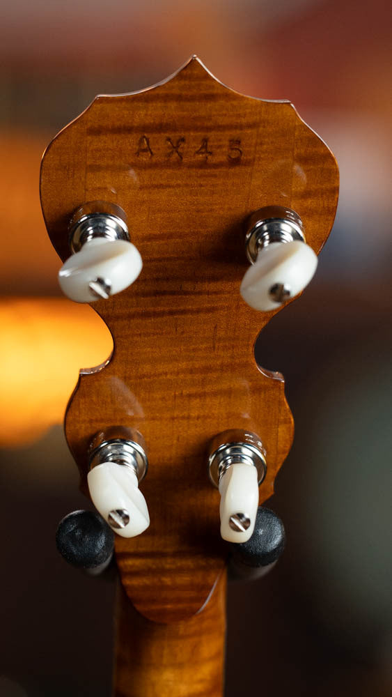 Close-up of the Deering Calico 5 String Curly Maple Resonator Banjo’s headstock by Deering Banjos, featuring four tuning pegs, code “AX45,” and polished curly maple wood with striking grain patterns.