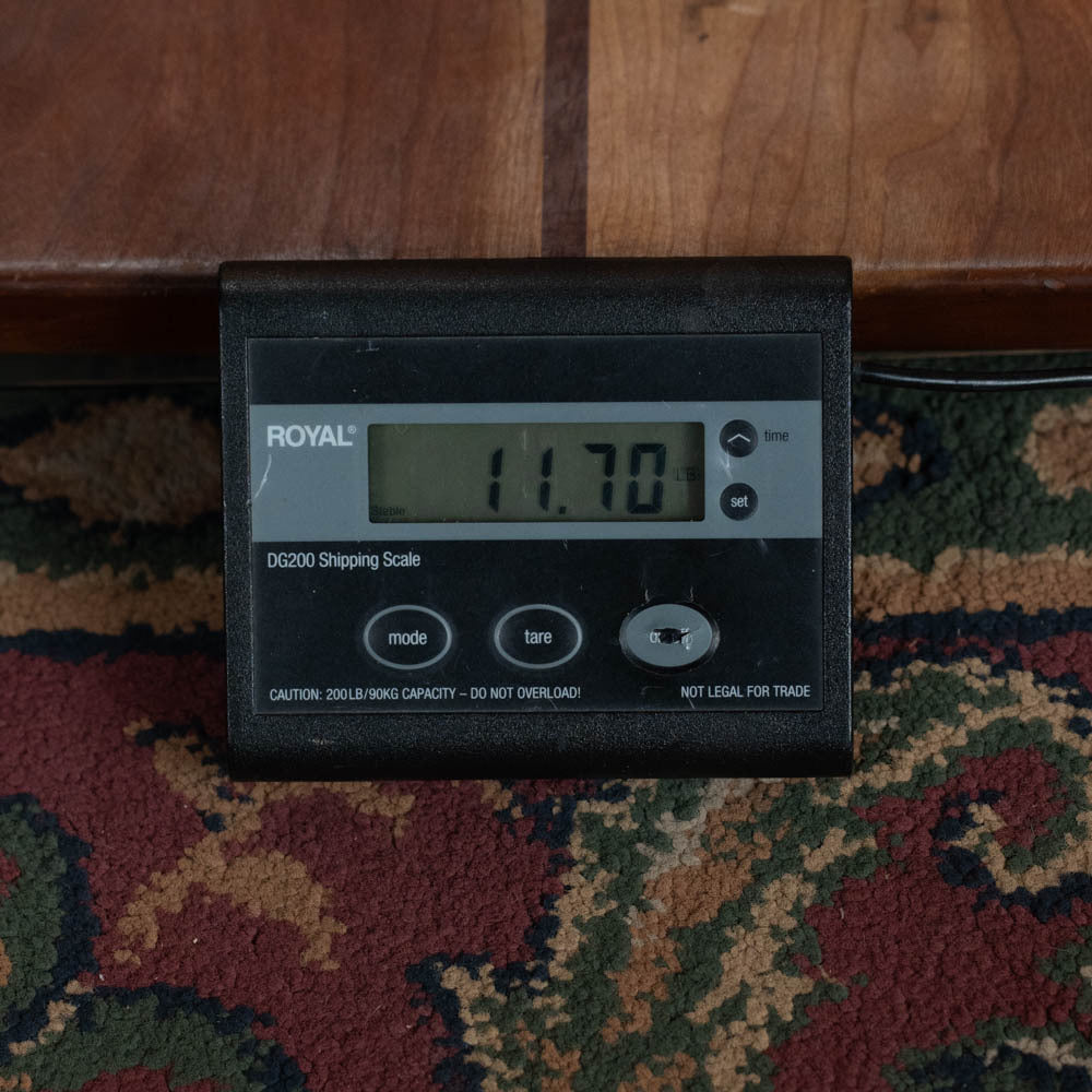 A digital shipping scale on a patterned carpet displays 11.70 pounds, with a Deering Banjos Calico 5 String Curly Maple Resonator Banjo w/Spikes Installed (#AX45) resting on top.