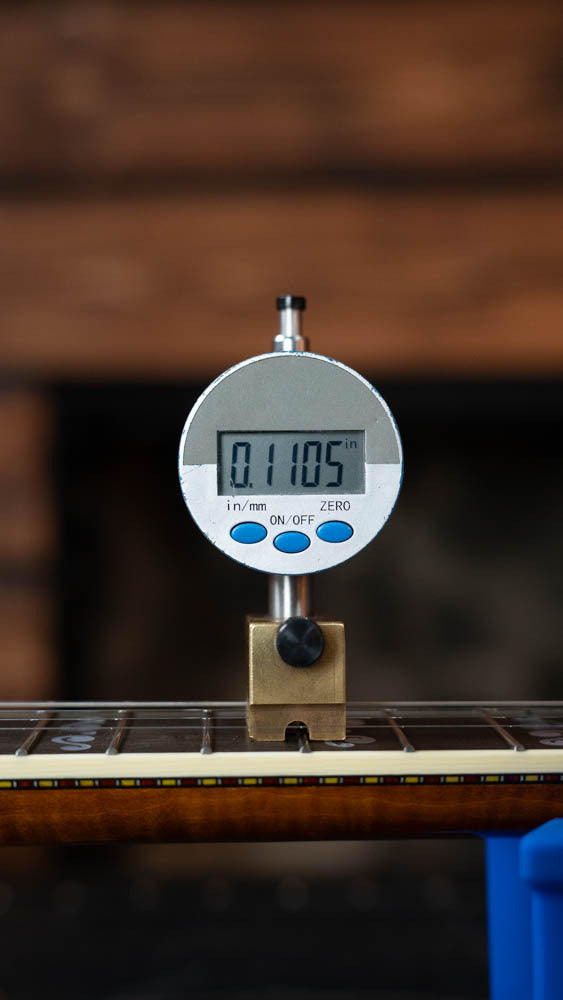 A digital gauge on a banjo fretboard shows 0.1105 inches of string height—just like adjusting a Deering Banjos Calico 5 String Curly Maple Resonator Banjo w/Spikes Installed #AX45 for professional playability.