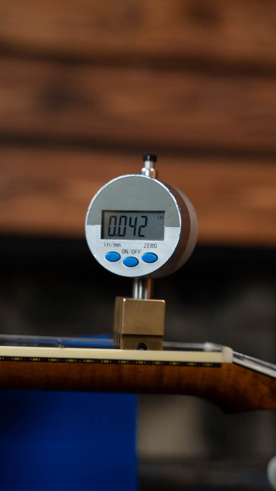 A digital gauge shows 0.042 inches while measuring the gap above the fretboard of a Deering Calico 5 String Curly Maple Resonator Banjo w/Spikes Installed #AX45 by Deering Banjos, set against a backdrop of blurred wood tones.