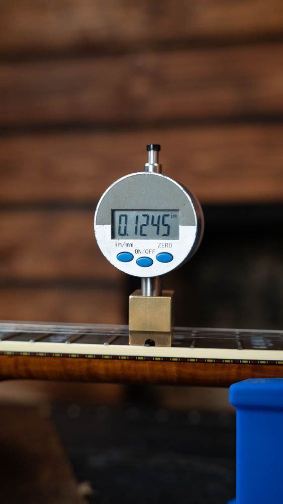 A digital gauge shows 0.1245 inches while measuring the height above the frets on a Deering Banjos Calico 5 String Curly Maple Resonator Banjo w/Spikes Installed #AX45, set against a blurred wooden background.