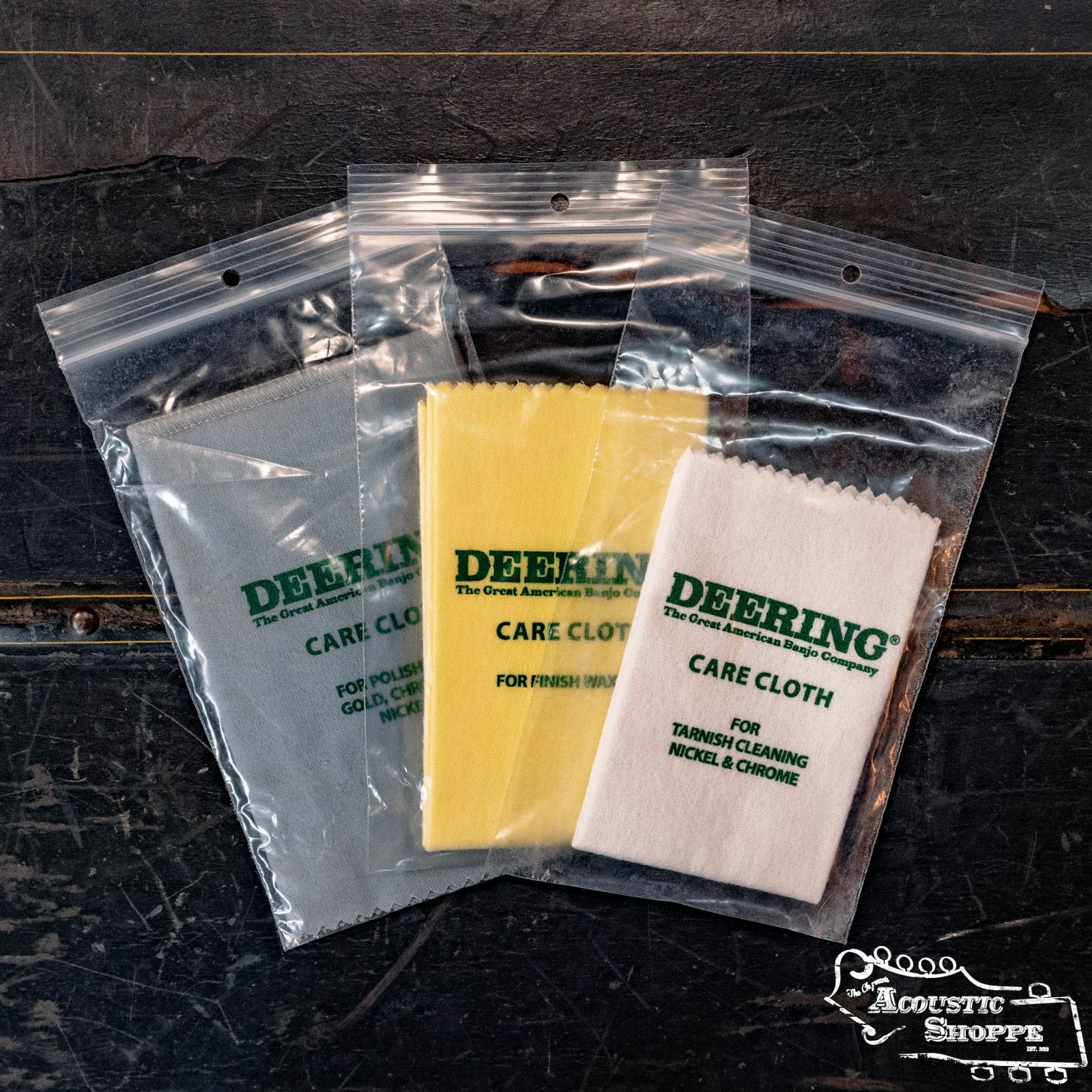 A Deering Banjos Care Cloth - Bundle of 3 (CC-SET) is shown in gray, yellow, and white—each individually packaged—on a dark wood surface with The Acoustic Shoppe logo in the lower right.