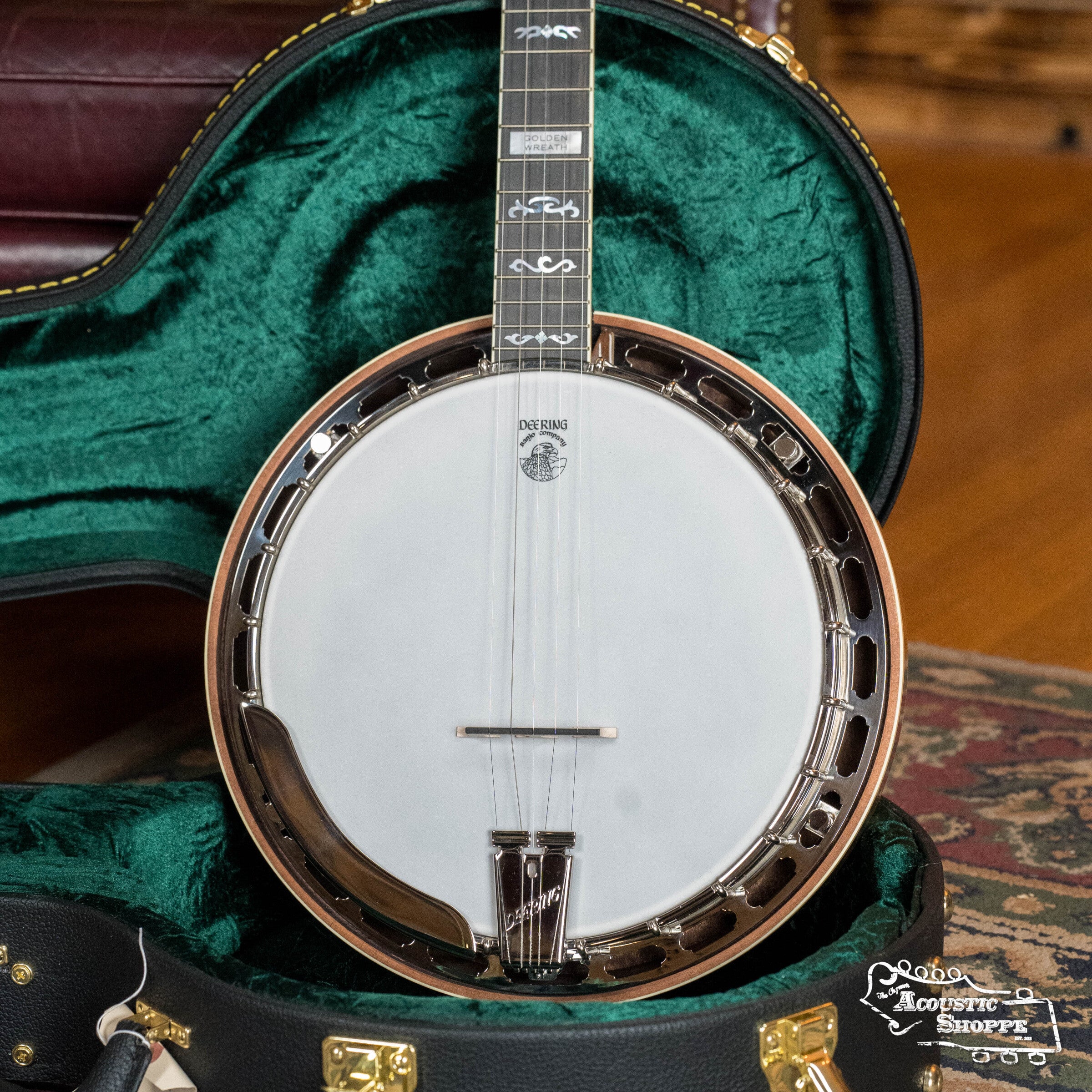 A close-up of the Deering Golden Wreath 5 String Resonator Banjo #AU47 by Deering Banjos, featuring a white drumhead, decorative fretboard inlays, and shiny metal rim inside a green velvet-lined hard case on a patterned rug.