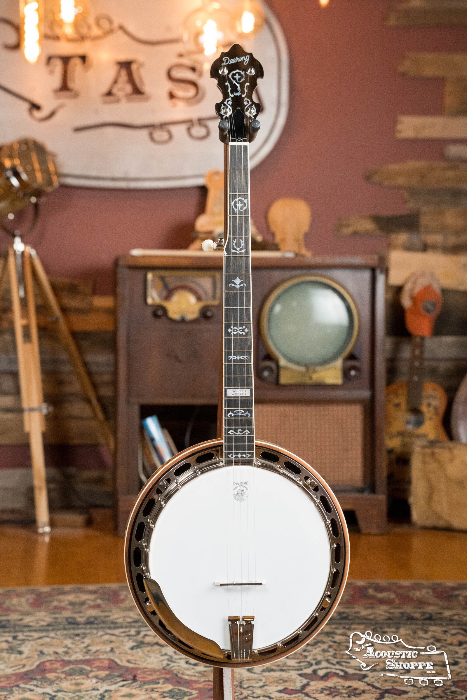 A Deering Golden Wreath 5 String Resonator Banjo #AU47 by Deering Banjos, featuring intricate inlays, stands upright on a stand in a cozy, vintage-style room with wooden furniture, a retro radio, and musical decor.