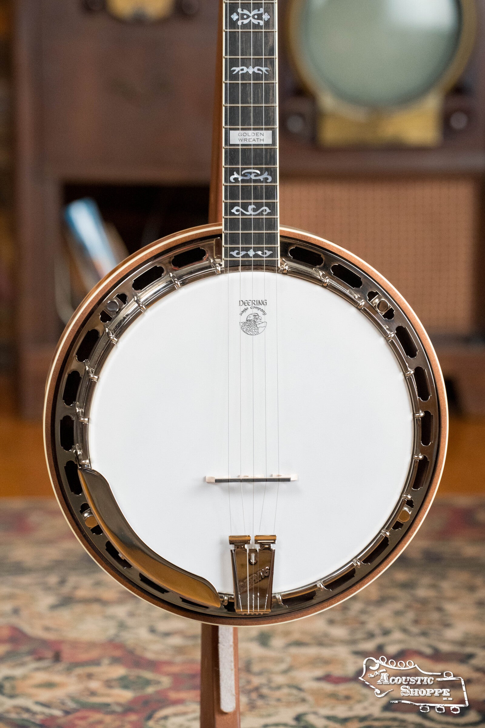 A close-up of the Deering Banjos Golden Wreath 5 String Resonator Banjo #AU47 on a stand shows its white drum head, metal rim, strings, and ornate neck inlays. The backdrop includes a patterned rug and vintage furniture.