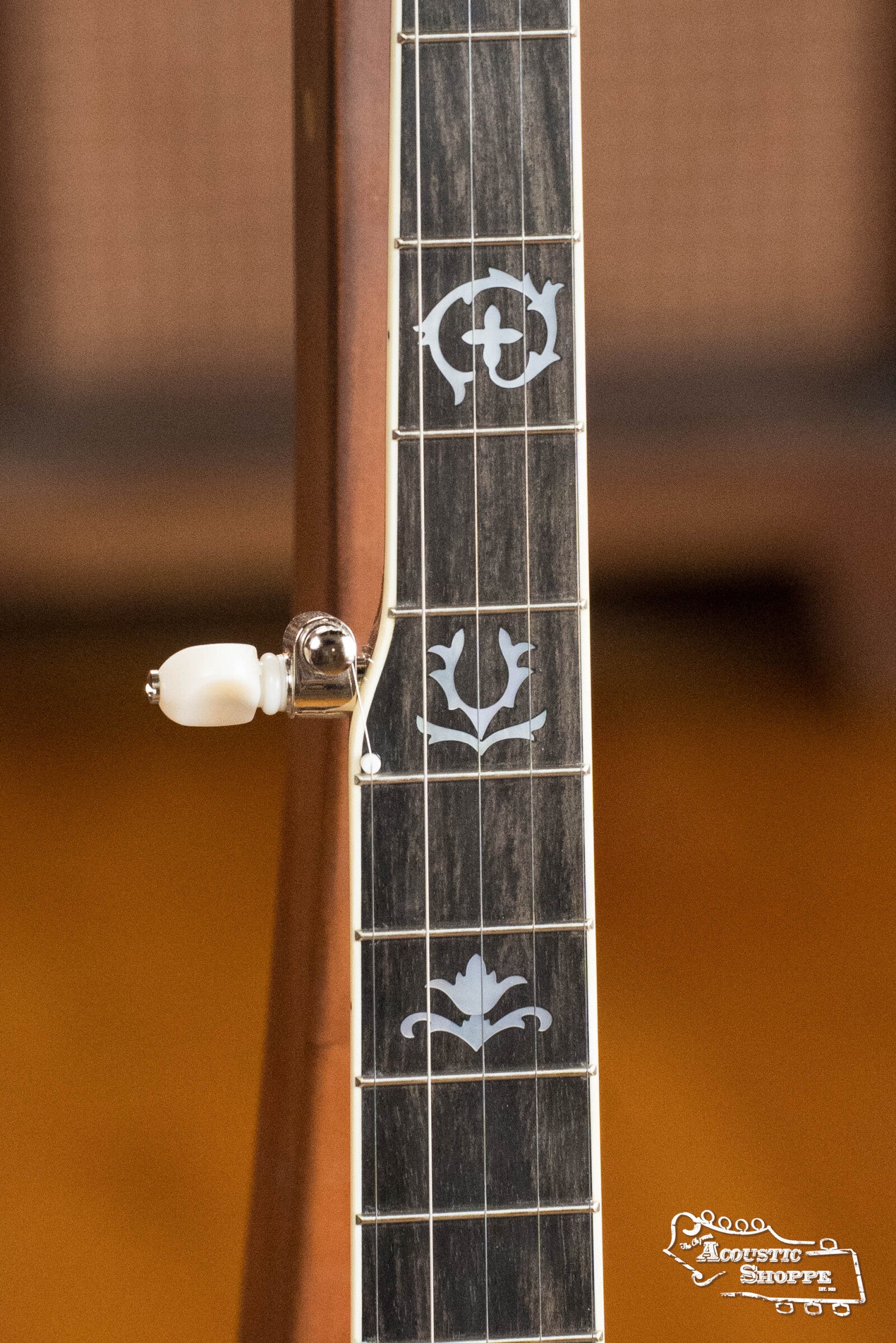 Close-up of the Deering Golden Wreath 5 String Resonator Banjo #AU47 by Deering Banjos, showing detailed white inlays on a dark fingerboard and a white tuning peg. The blurred background features Acoustic Shoppe in the bottom right.