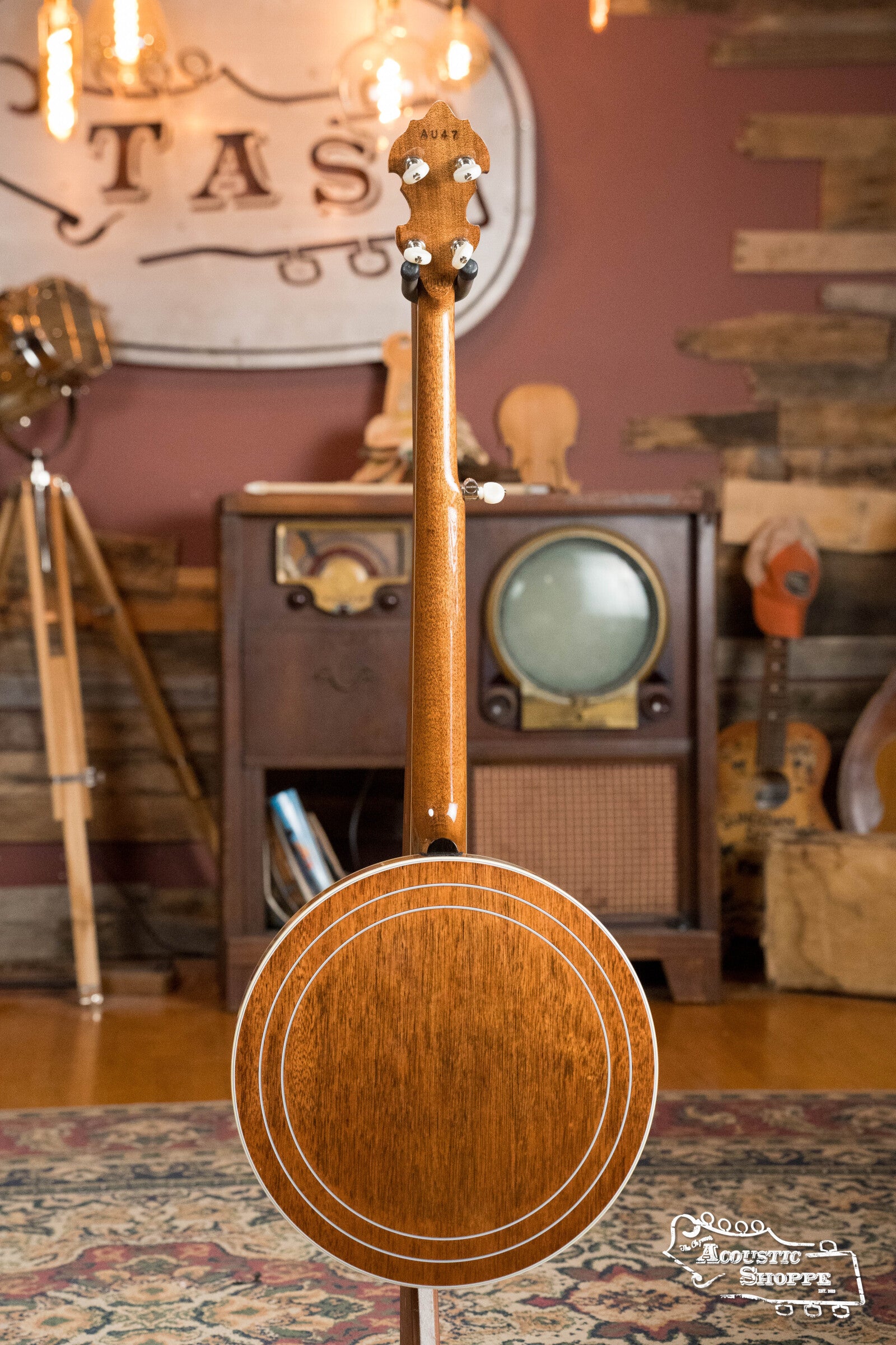 The Deering Golden Wreath 5 String Resonator Banjo #AU47 by Deering Banjos is shown upright from the back, highlighting its wooden finish and white inlay rings in a retro room with vintage TV, radio, camera, and rustic wood walls.