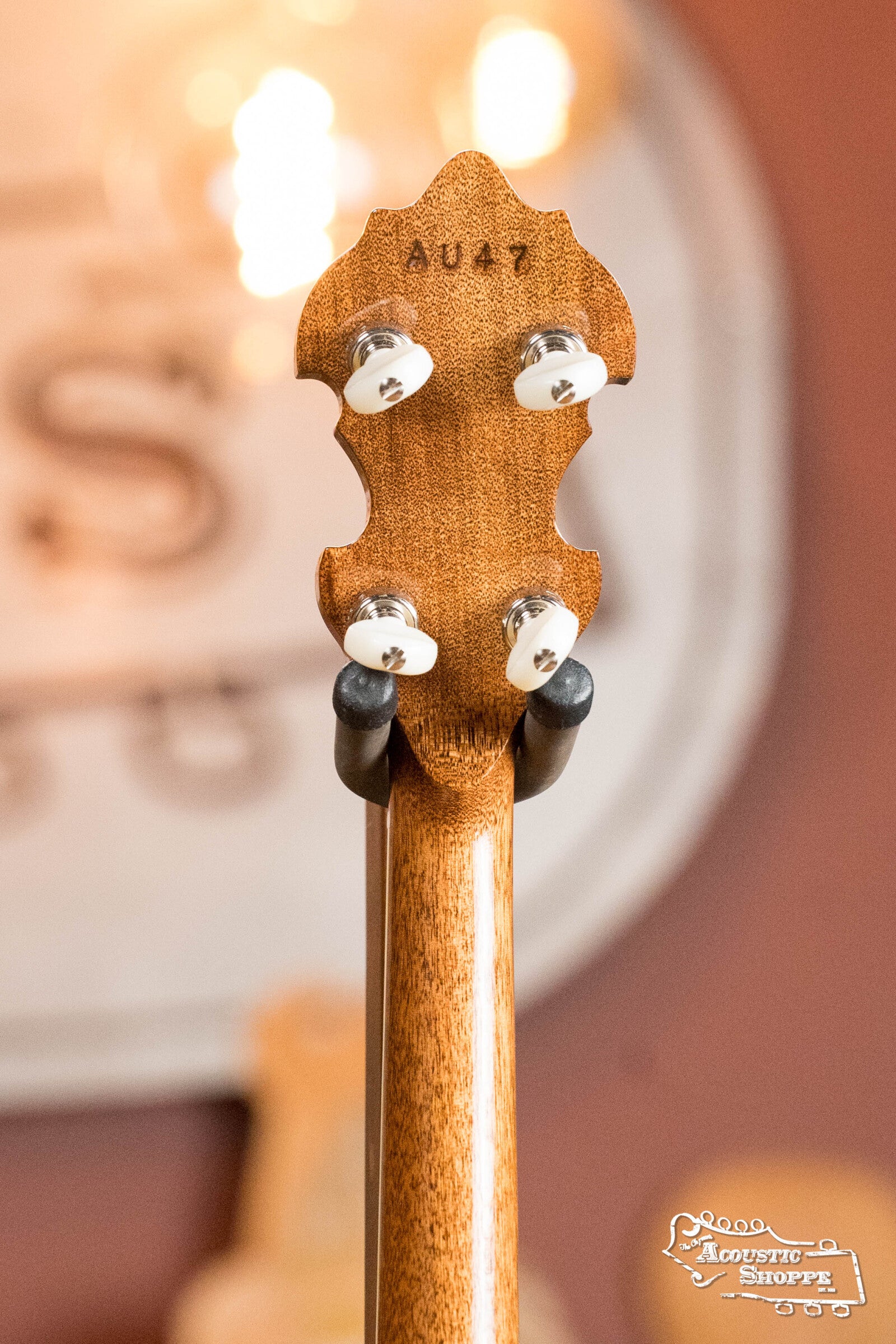 Close-up of the Deering Golden Wreath 5 String Resonator Banjo #AU47 headstock by Deering Banjos, showing four tuning pegs and AU47 engraving on the glowing natural wood, under warm light with a blurred background and a white sign visible.