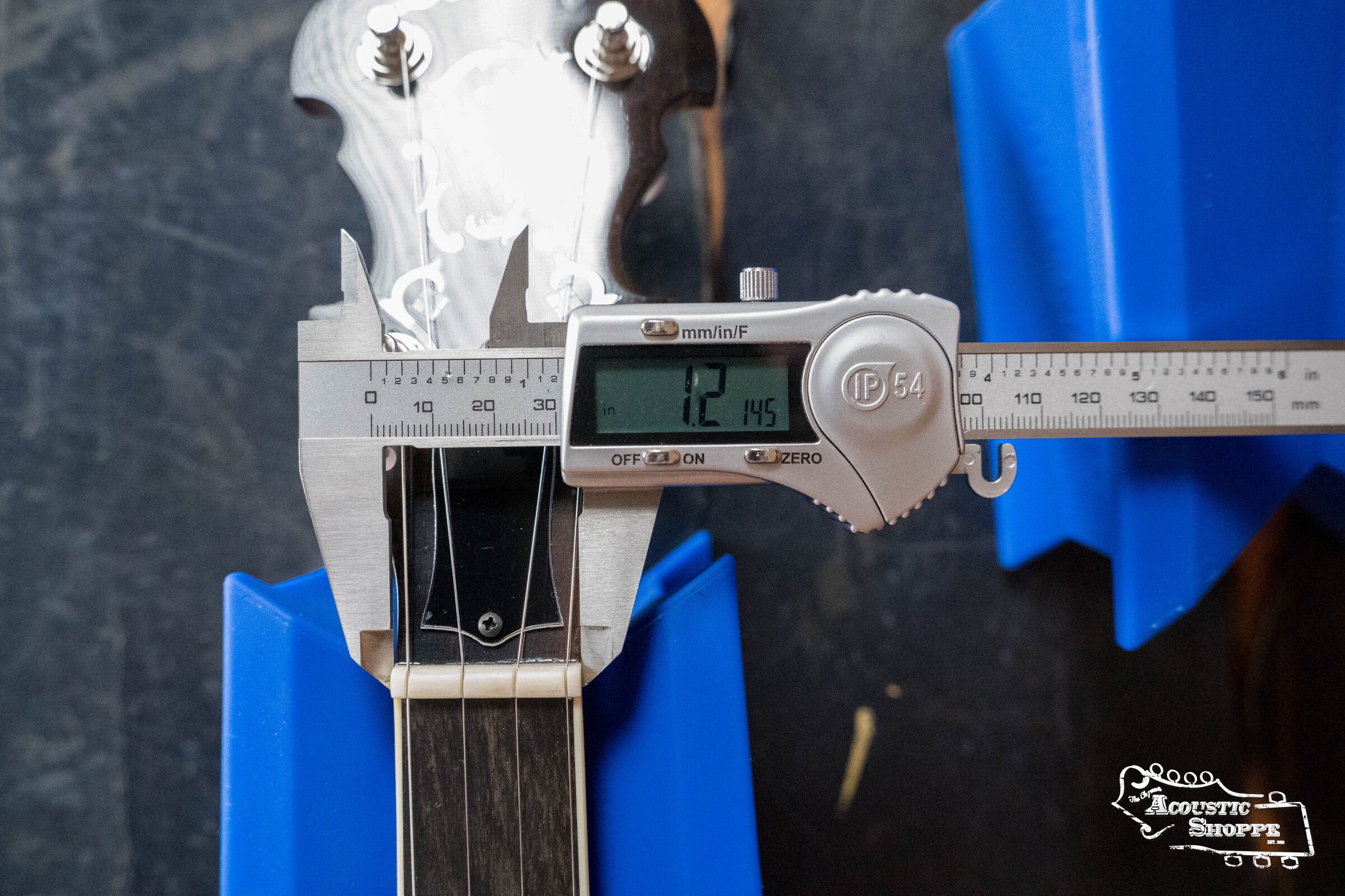A digital caliper measures a guitar neck at 1.2145 inches, with the guitar in blue holders. The Acoustic Shoppe logo appears bottom right, showcasing their Deering Banjos Deering Golden Wreath 5 String Resonator Banjo #AU47 selection.