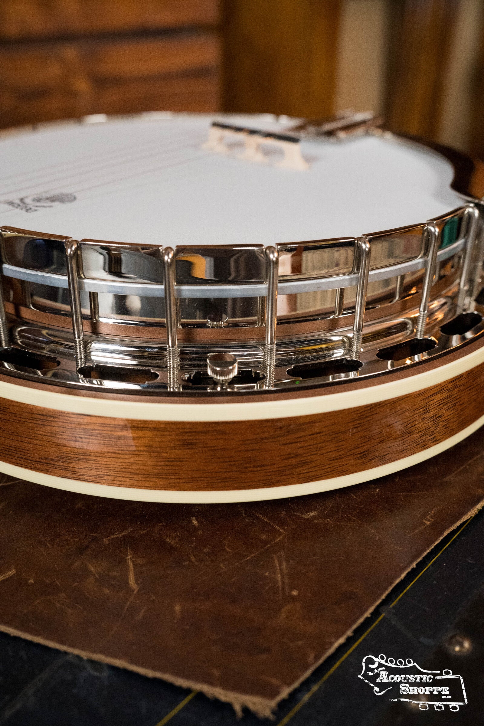 A close-up of the Deering Golden Wreath 5 String Resonator Banjo #AU47 by Deering Banjos rests on leather, showcasing its wooden body, metal resonator, white drumhead with strings, and The Acoustic Shoppe logo in the corner.