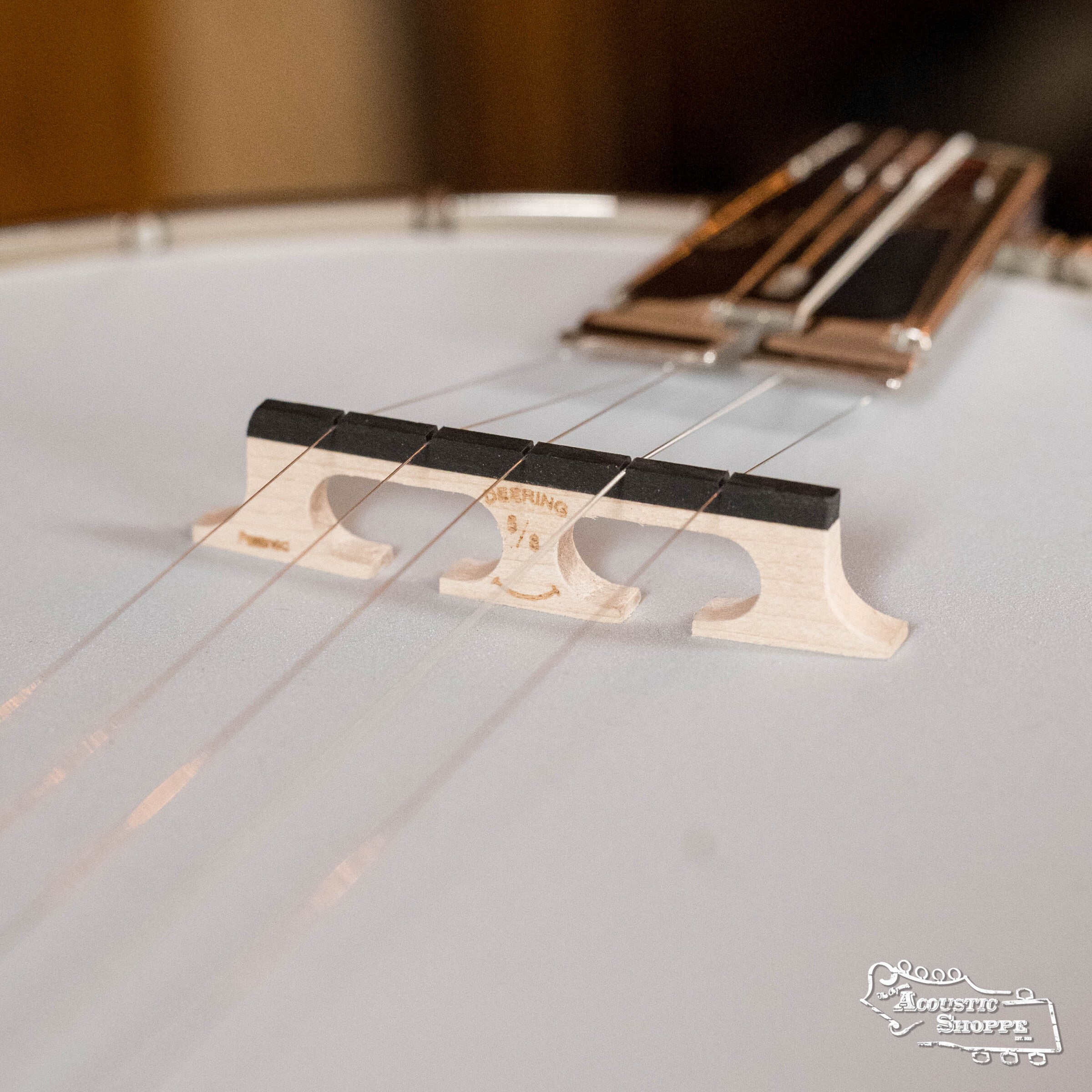 Close-up of a Deering Golden Wreath 5 String Resonator Banjo #AU47 bridge by Deering Banjos, with five strings over a white drumhead. The Acoustic Shoppe appears in the bottom right corner.
