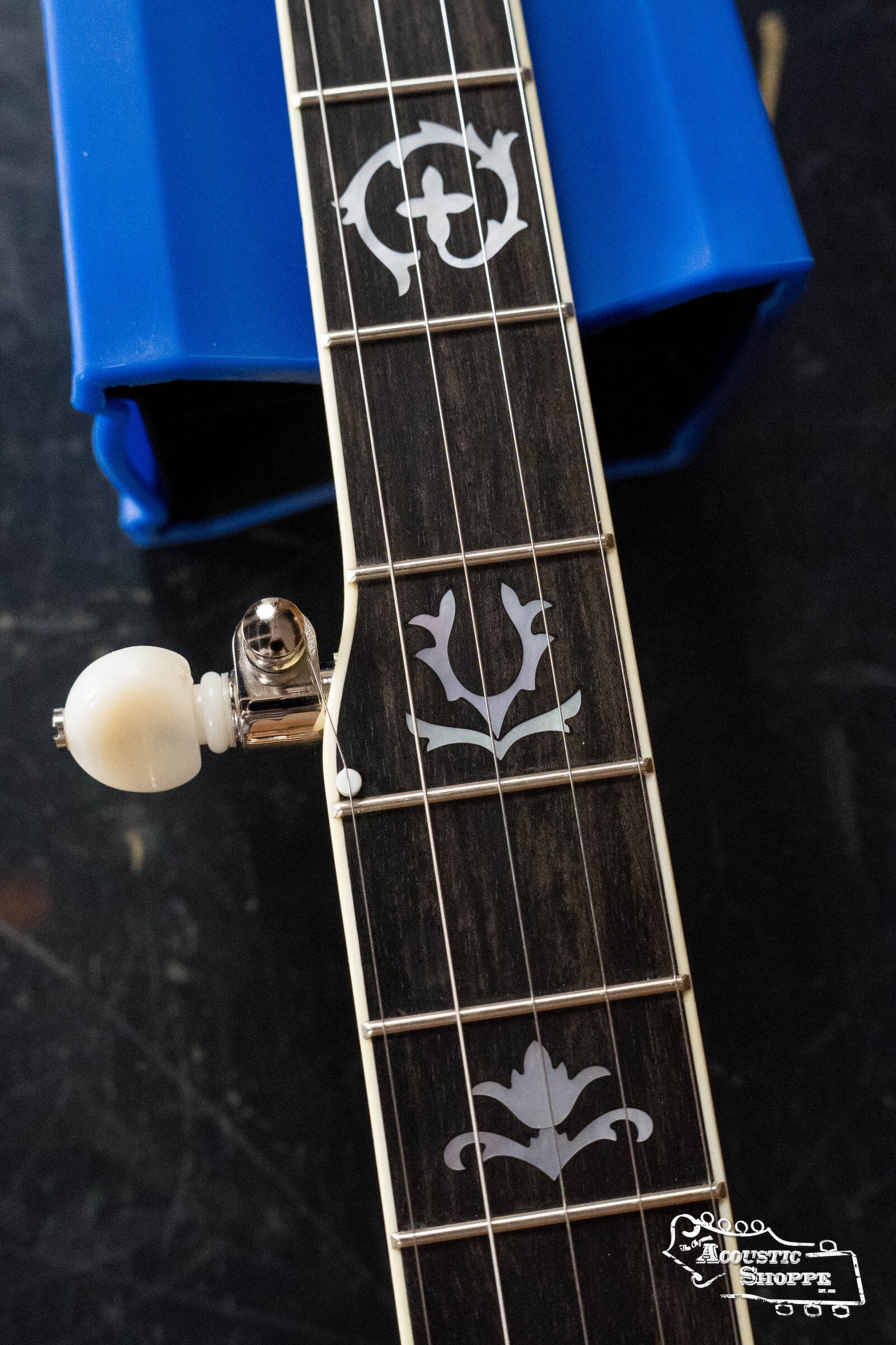 A close-up of the Deering Banjos Golden Wreath 5 String Resonator Banjo #AU47s neck shows decorative fretboard inlays and a headstock tuning peg, beside a blue object. The Acoustic Shoppe logo is in the bottom right corner.