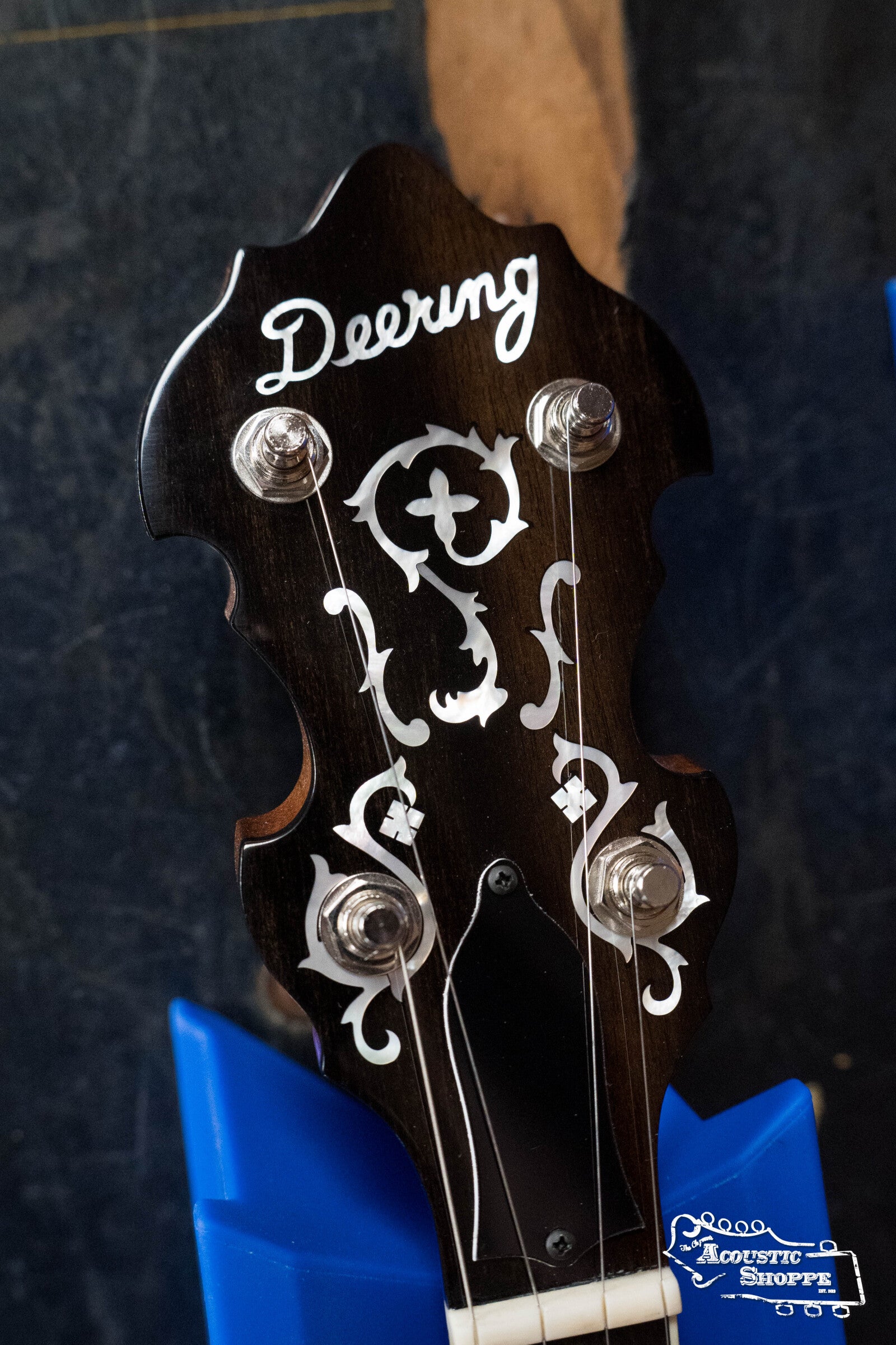 Close-up of a Deering Banjos Golden Wreath 5 String Resonator Banjo #AU47 headstock, featuring intricate white inlays and displayed on a blue stand. The Acoustic Shoppe logo appears in the corner—a true bluegrass classic.