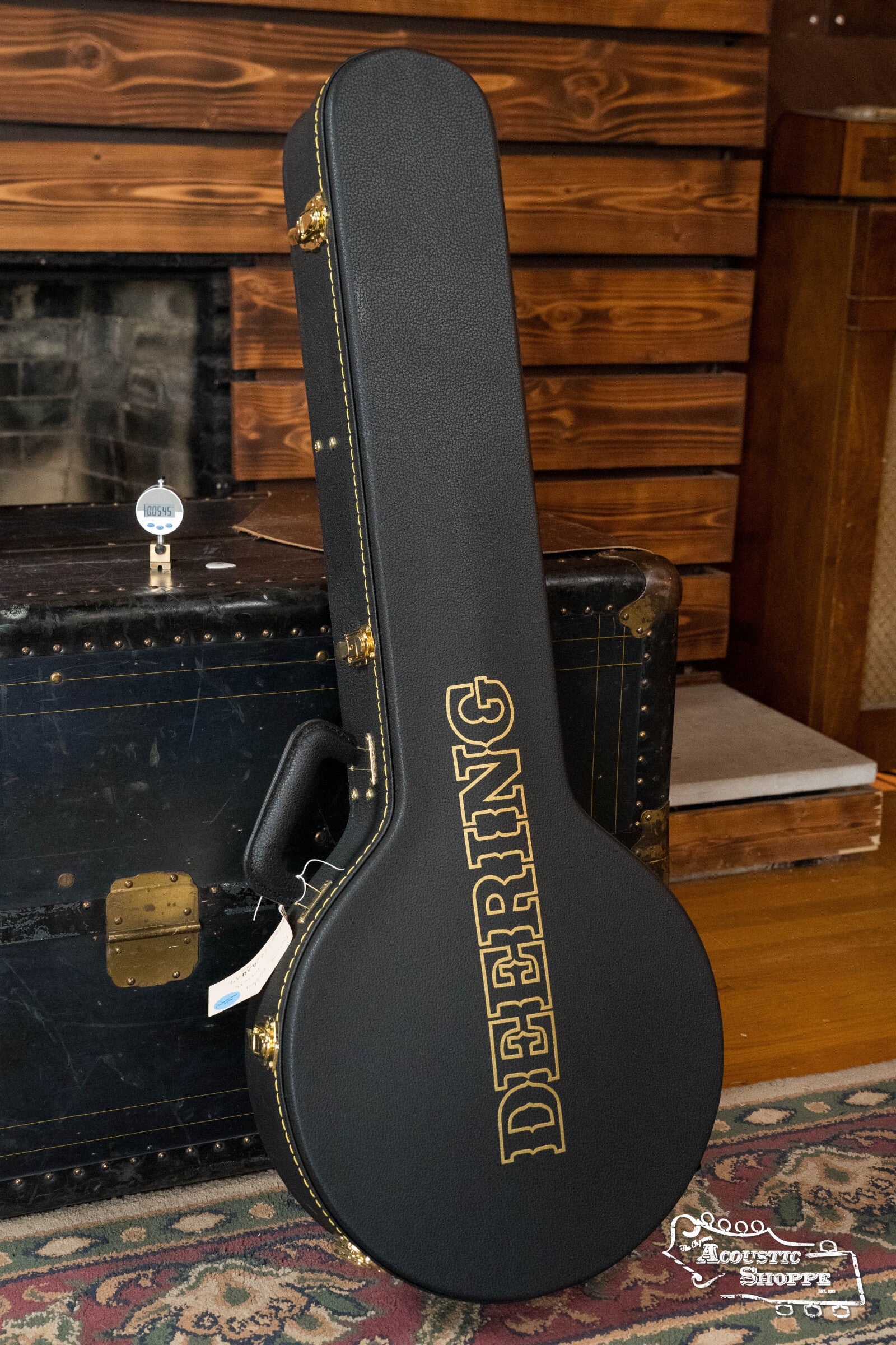 A black Deering Banjos case with gold lettering stands upright on a vintage trunk in a cozy, wood-paneled room—the perfect showcase for your Deering Golden Wreath 5 String Resonator Banjo #AU47.
