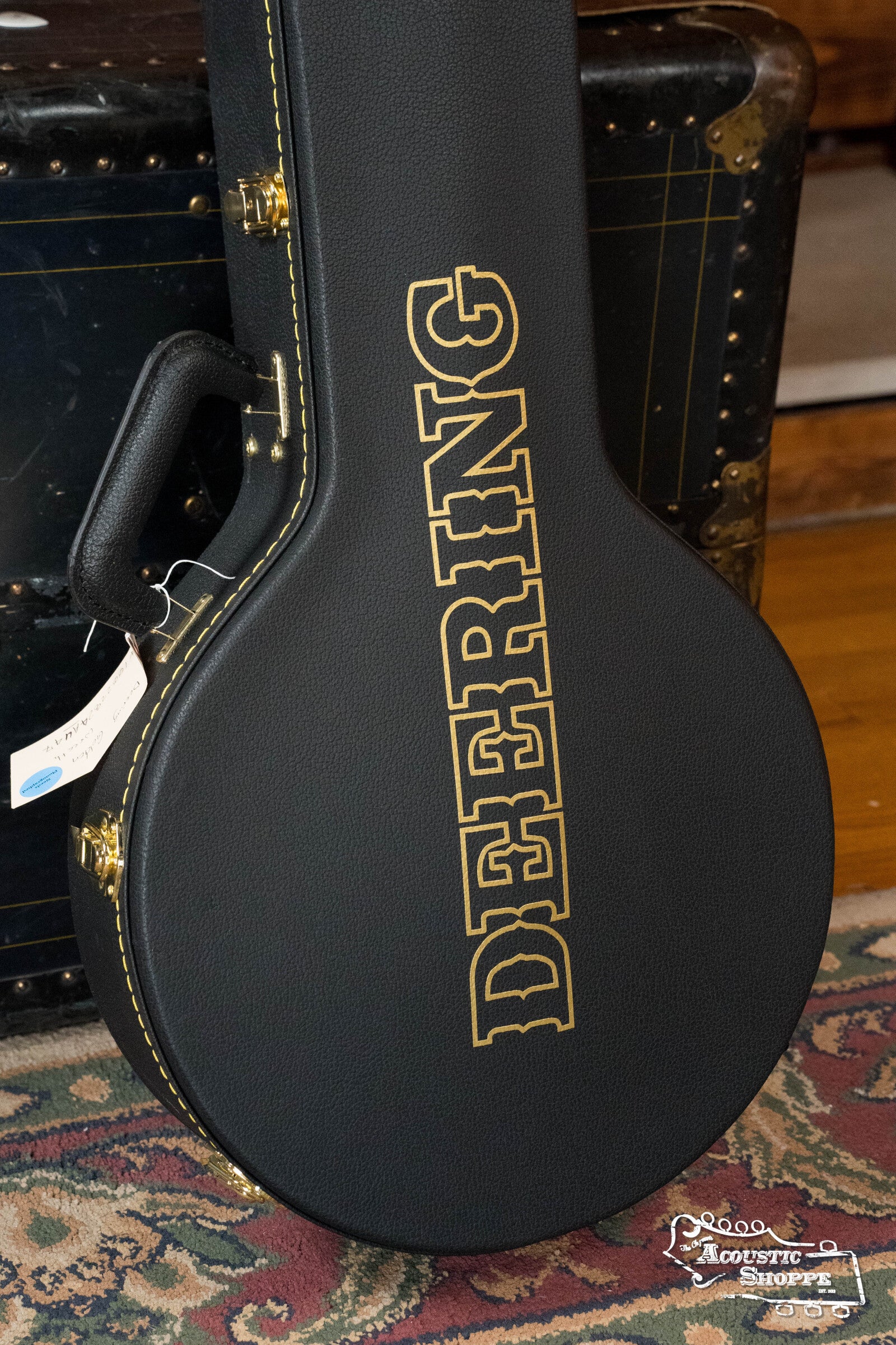A black Deering Banjos hard case with gold lettering stands on a patterned rug by a vintage trunk. A white tag hangs from the handle, and the Acoustic Shoppe 2015 logo is visible. Ideal for the Deering Golden Wreath 5 String Resonator Banjo #AU47.