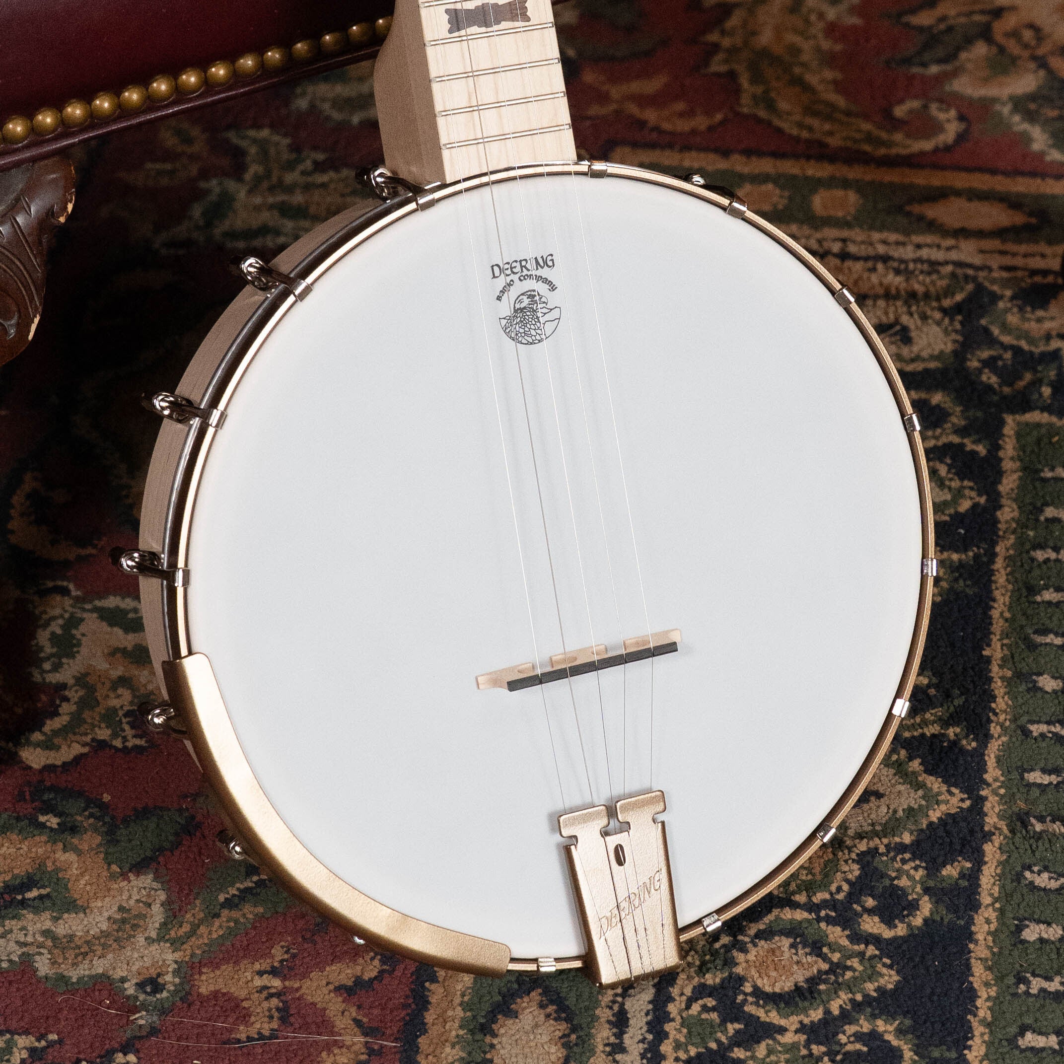 A close-up of the Deering Goodtime 5 String Open Back Banjo w/Spikes Installed #2513 by Deering Banjos, featuring a maple rim, white drumhead, strings, and metal bridge as it rests on an ornate patterned rug.