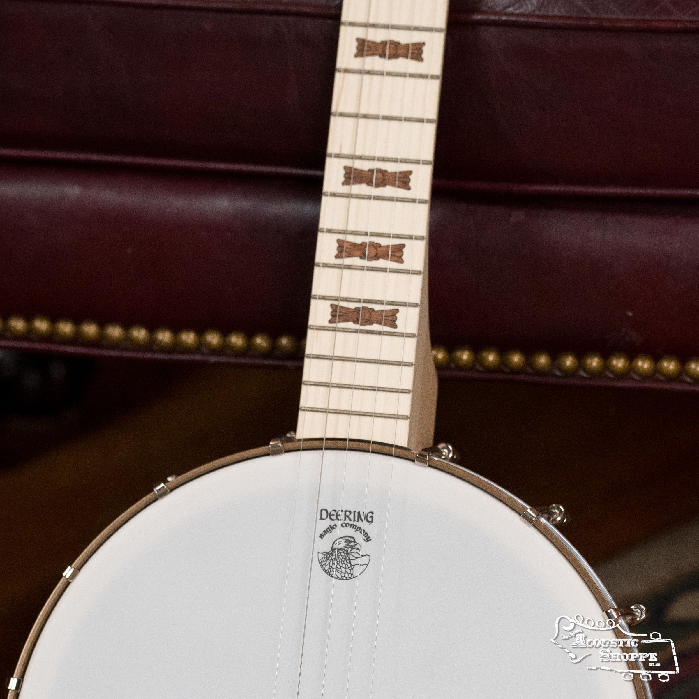 A close-up of a Deering Banjos Goodtime Deco 5-String Banjo #52407’s neck and head shows distinctive fretboard inlays and the Deering logo, with part of a dark red leather chair visible in the background.