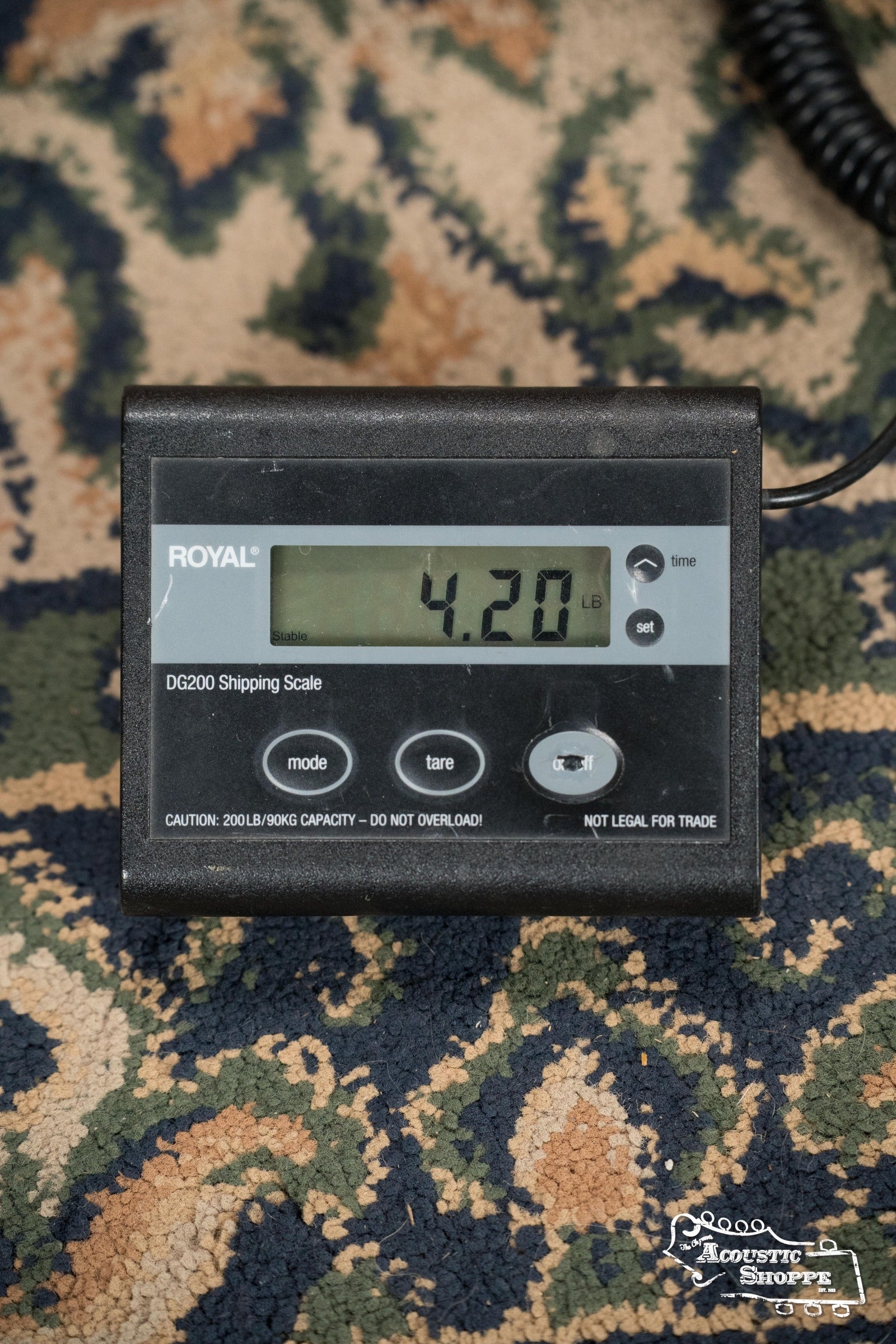 A digital shipping scale reads 4.20 lbs, likely weighing a Deering Banjos Goodtime Deco 5-String Banjo (#52407). The scale has mode, tare, and on/off buttons, a max capacity warning, and rests on patterned carpet.