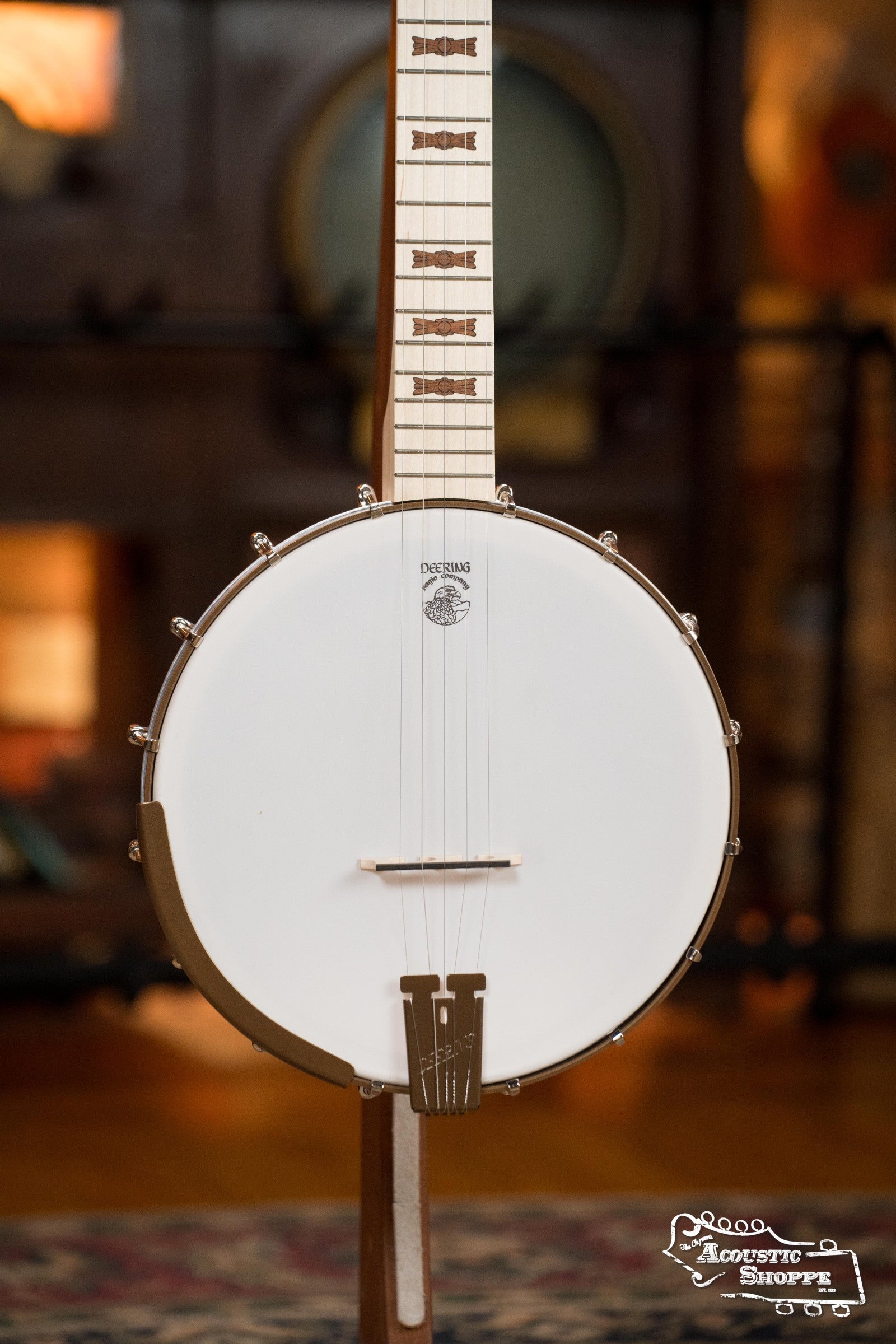 A close-up of the Deering Goodtime Deco 5-String Banjo #52407 by Deering Banjos stands upright, displaying its white drumhead, light neck with decorative inlays, and metal tailpiece against a softly blurred background.