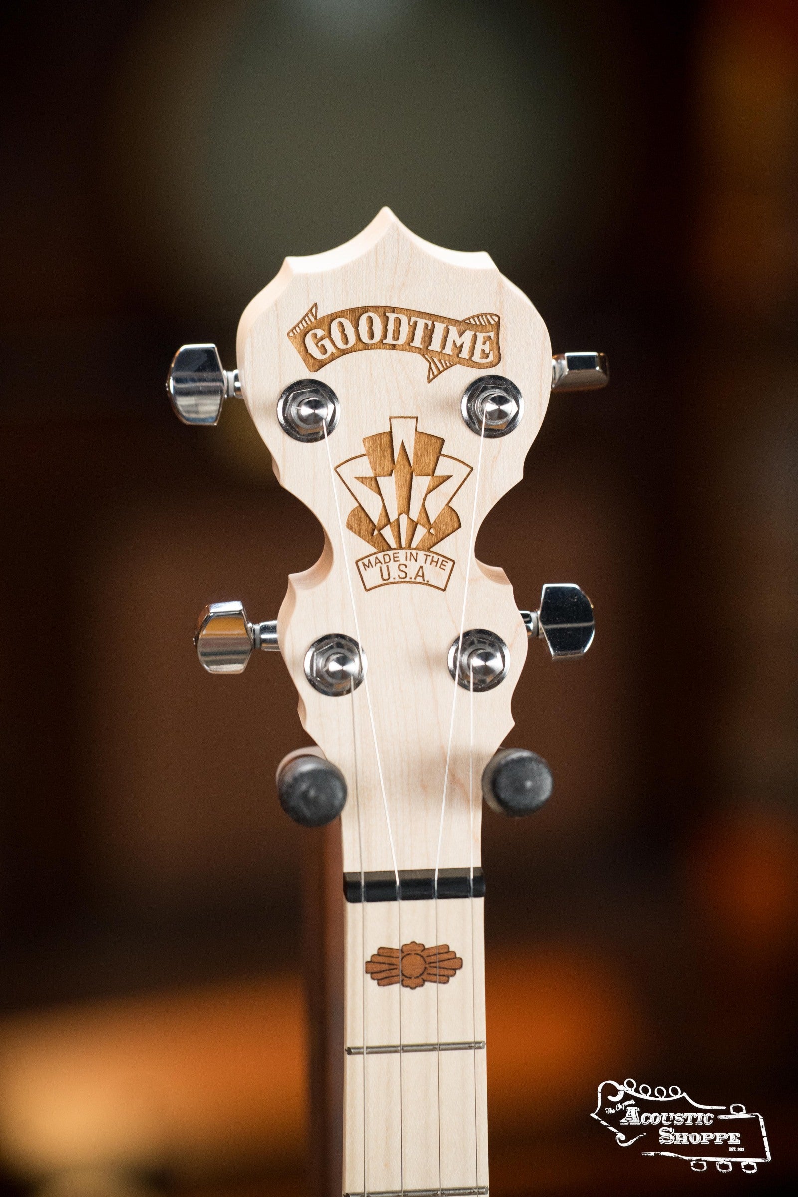 Close-up of the Deering Goodtime Deco 5-String Banjo #52407 headstock by Deering Banjos, featuring GOODTIME and Made in U.S.A. engraved on light wood and four tuning pegs against a blurred brown background.