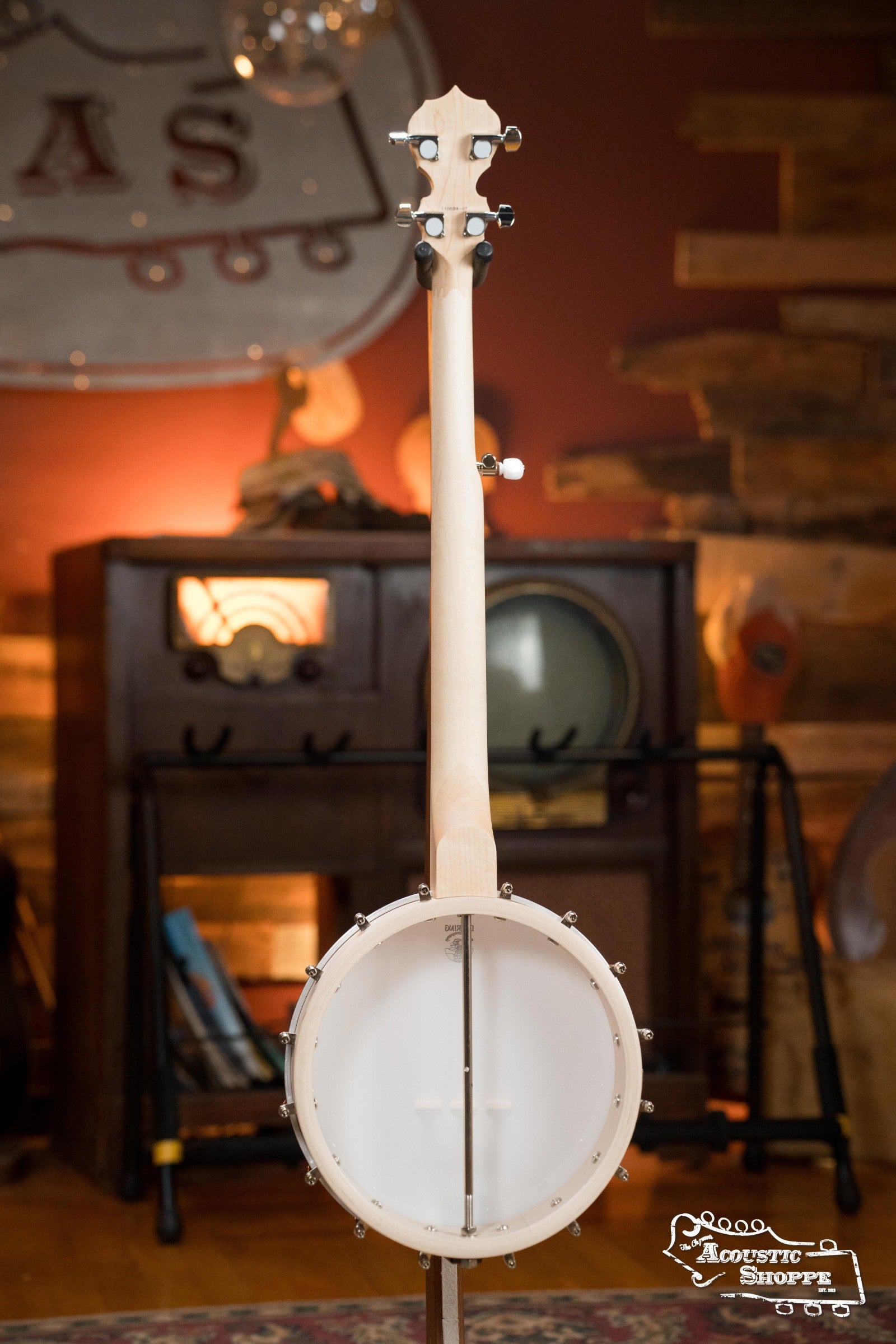 A Deering Banjos Goodtime Deco 5-String Banjo #52407 is displayed from the back, upright in a warmly lit room with wooden accents and vintage gear. The Acoustic Shoppe logo is visible in the bottom right corner.